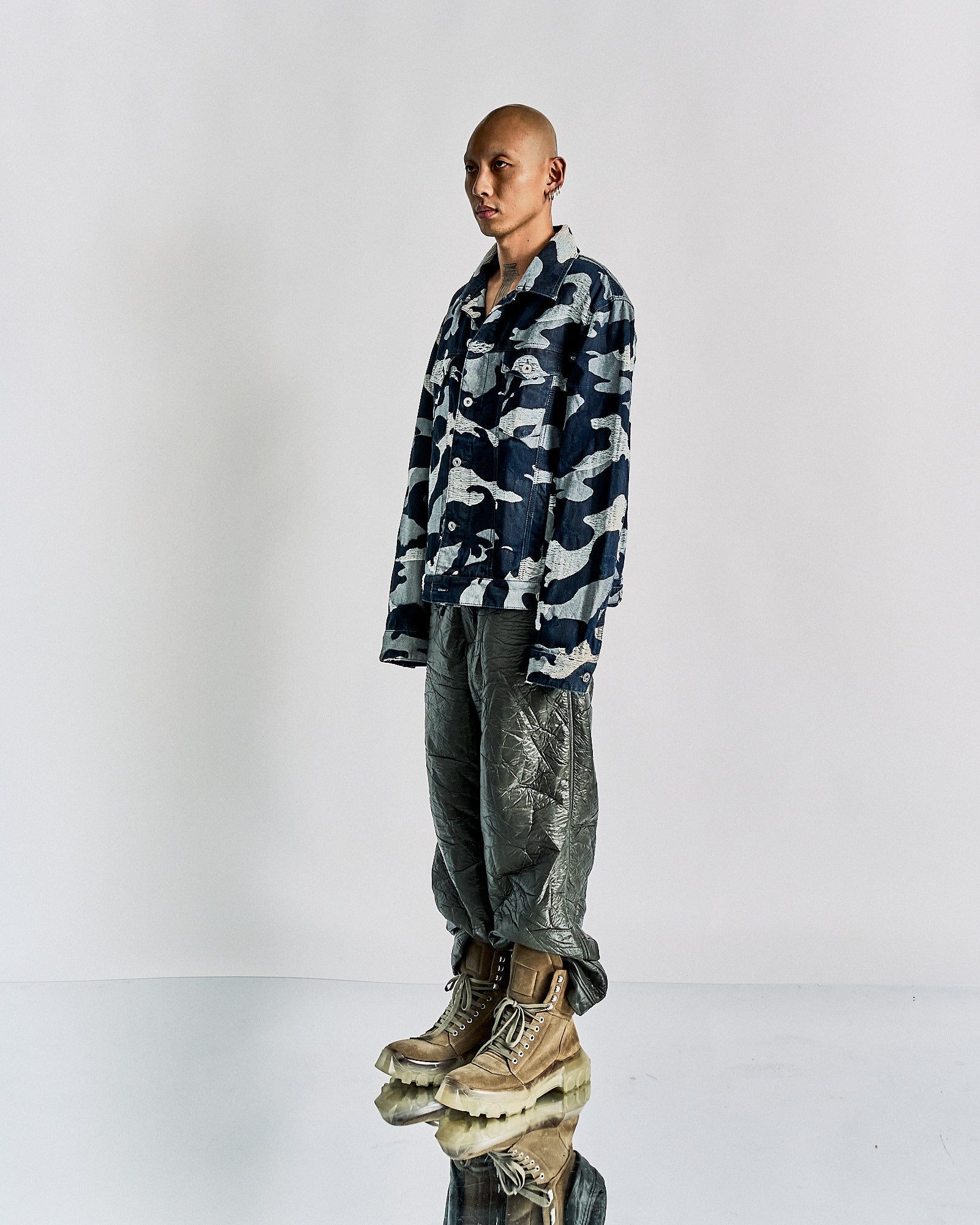 A person poses confidently in a blue camouflage Valentino jacket and metallic crinkled pants. They wear tan boots. The setting is minimal and modern.