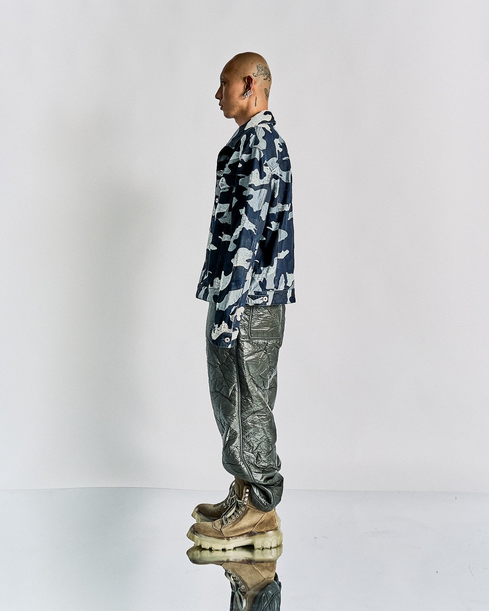 A person stands in profile against a plain backdrop, wearing a blue camouflage Valentino jacket and shiny metallic pants. They're on chunky, textured sandals, conveying a futuristic vibe.