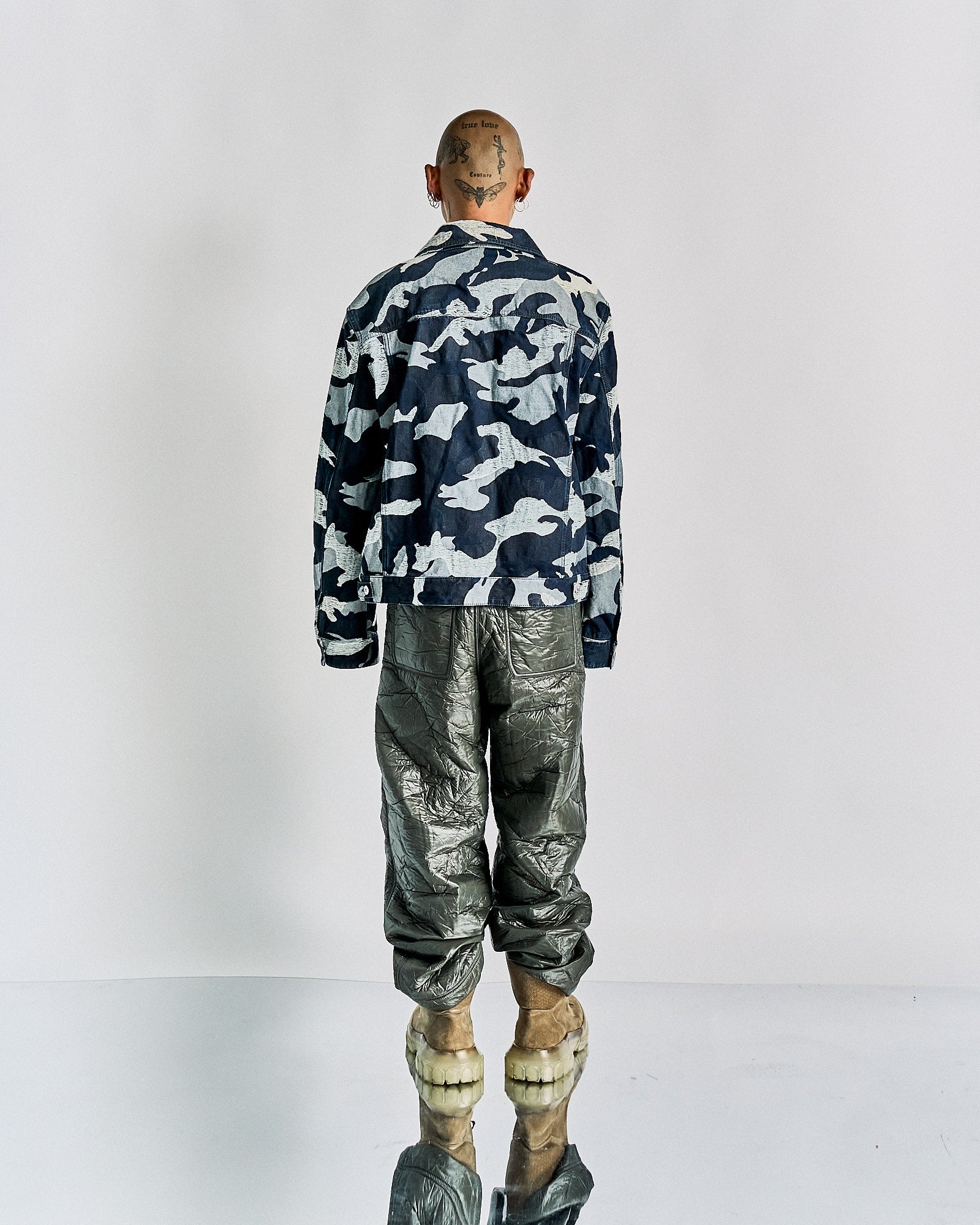 Back view of a person wearing a blue and white camouflage Valentino jacket, shiny metallic pants, and unique, chunky footwear. The head is tattooed.