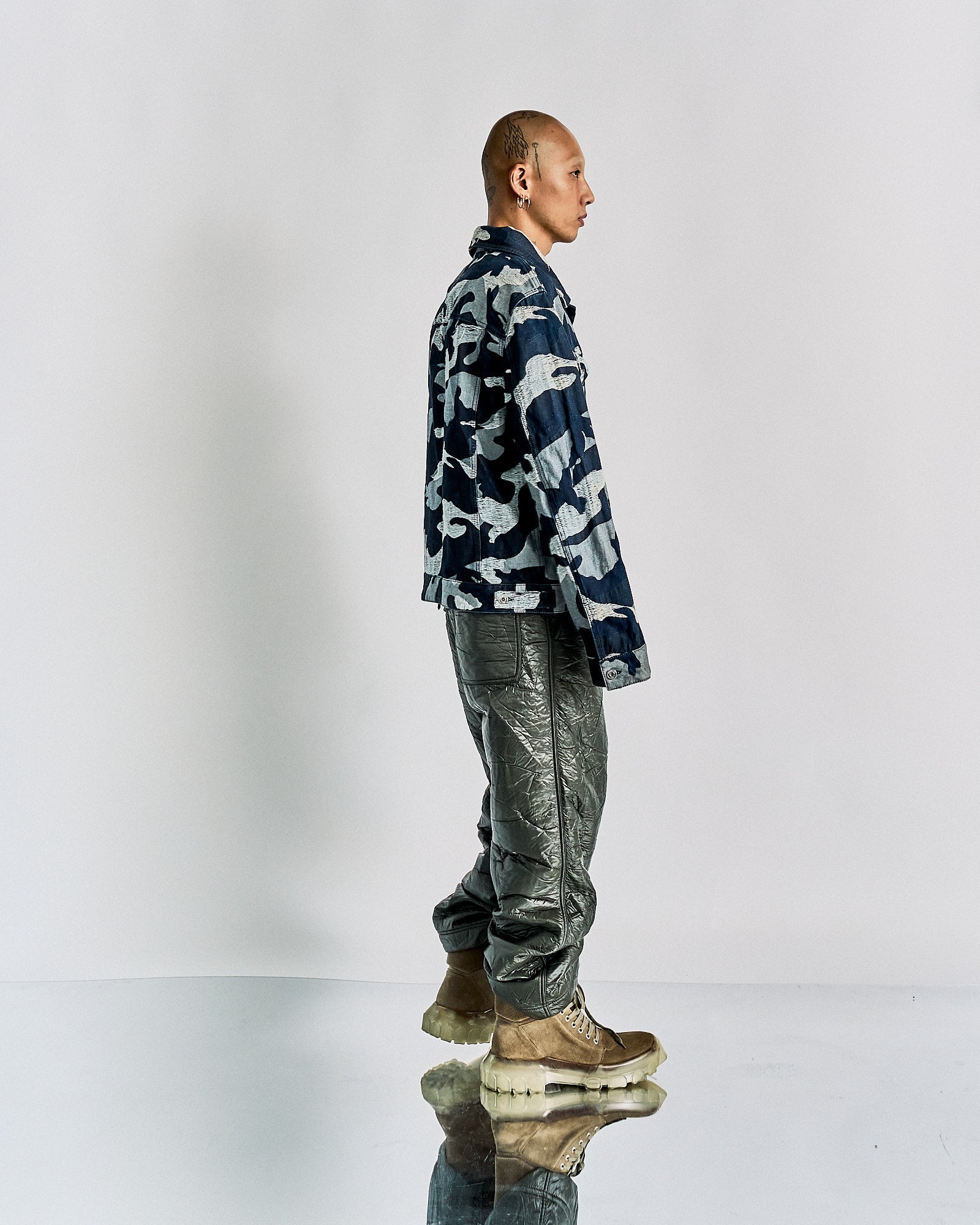 Man in side profile wearing a blue Valentino camo jacket, textured dark pants, and brown boots with clear soles. Background is plain white. Casual, fashion-focused vibe.