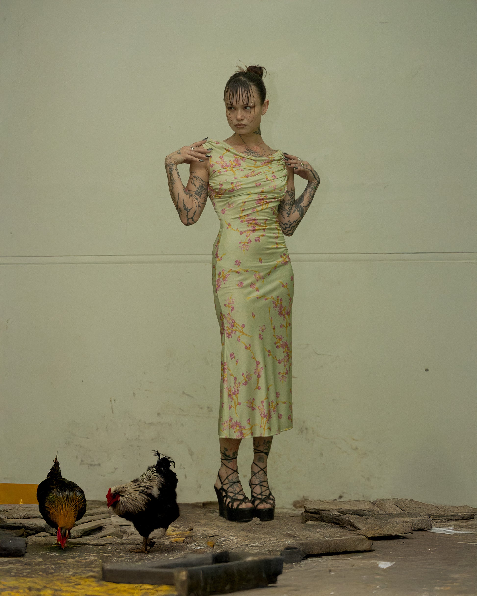 A woman standing on a textured surface wearing a floral sleeveless floor-length Versace dress with a side-zip and hook closure. There are two chickens near her feet.
