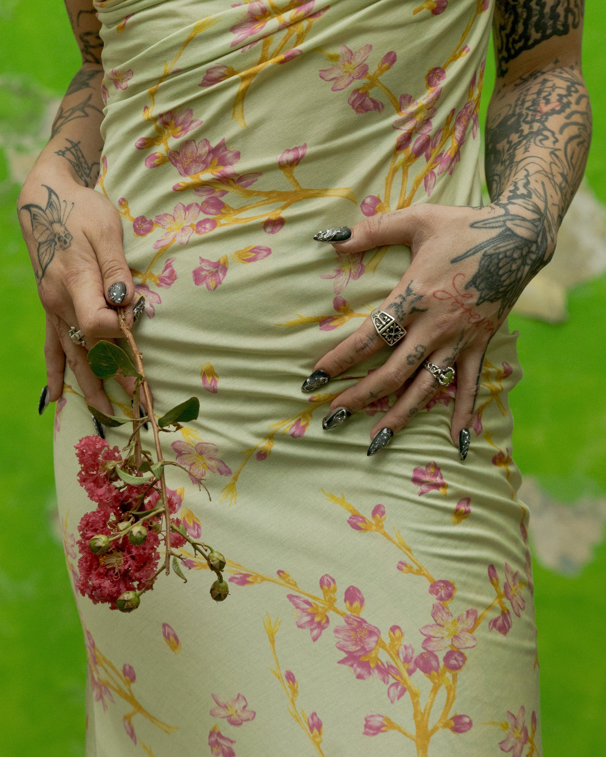 Person wearing a floral Versace dress with tattoos and rings on a green background.