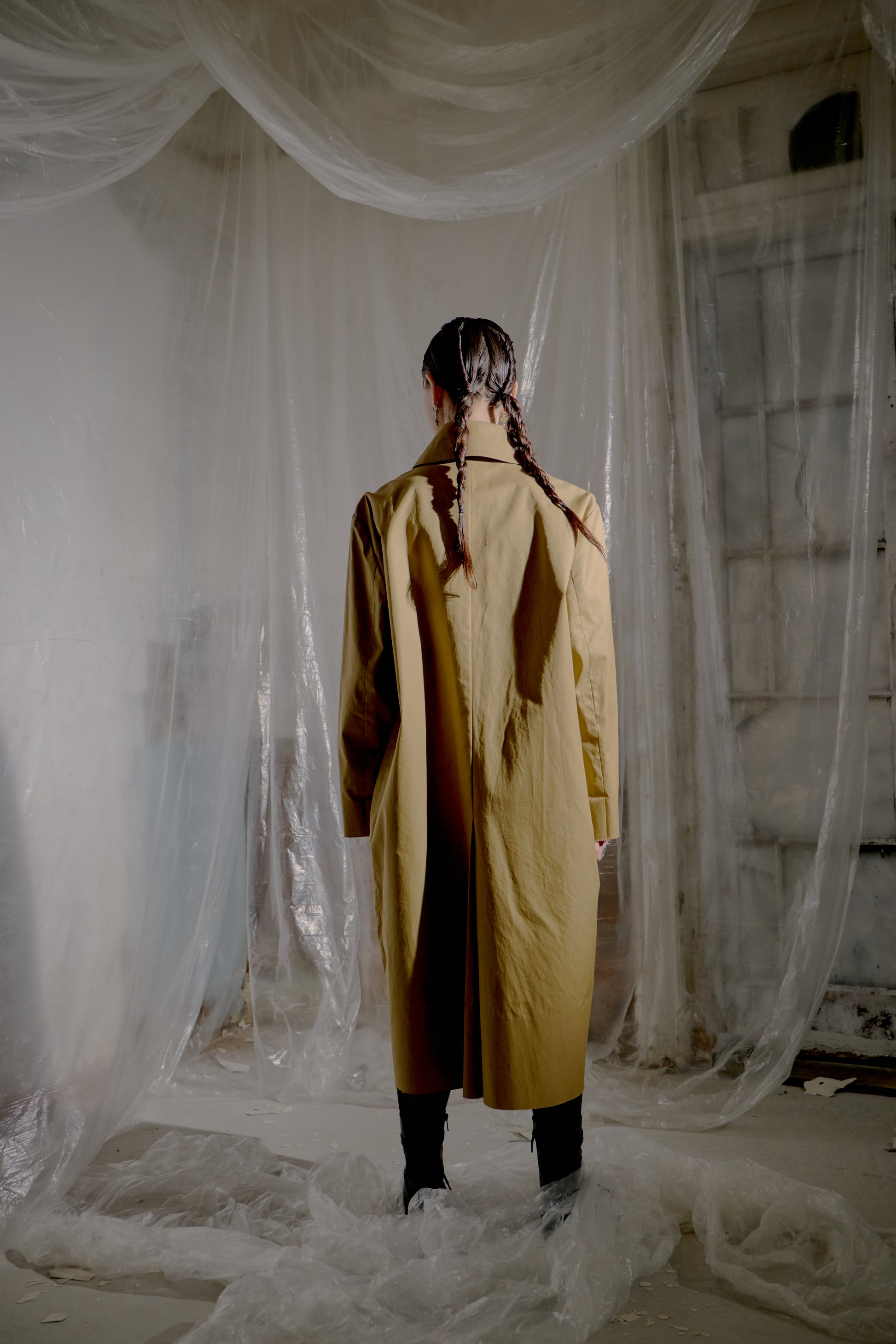 Person wearing a long beige Vetements coat standing in a room with sheer curtains.