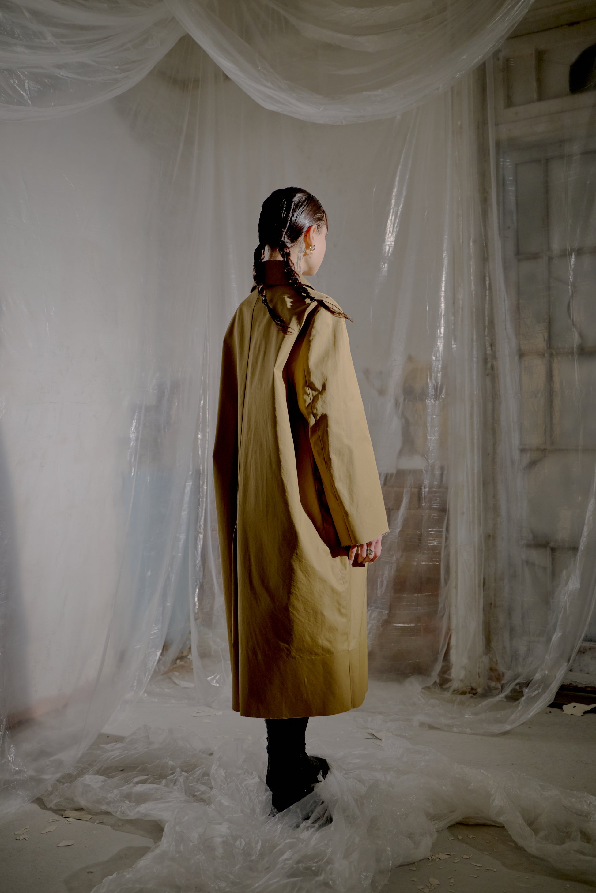 Person wearing a long beige Vetements coat standing in a room with white curtains and a rustic wall.