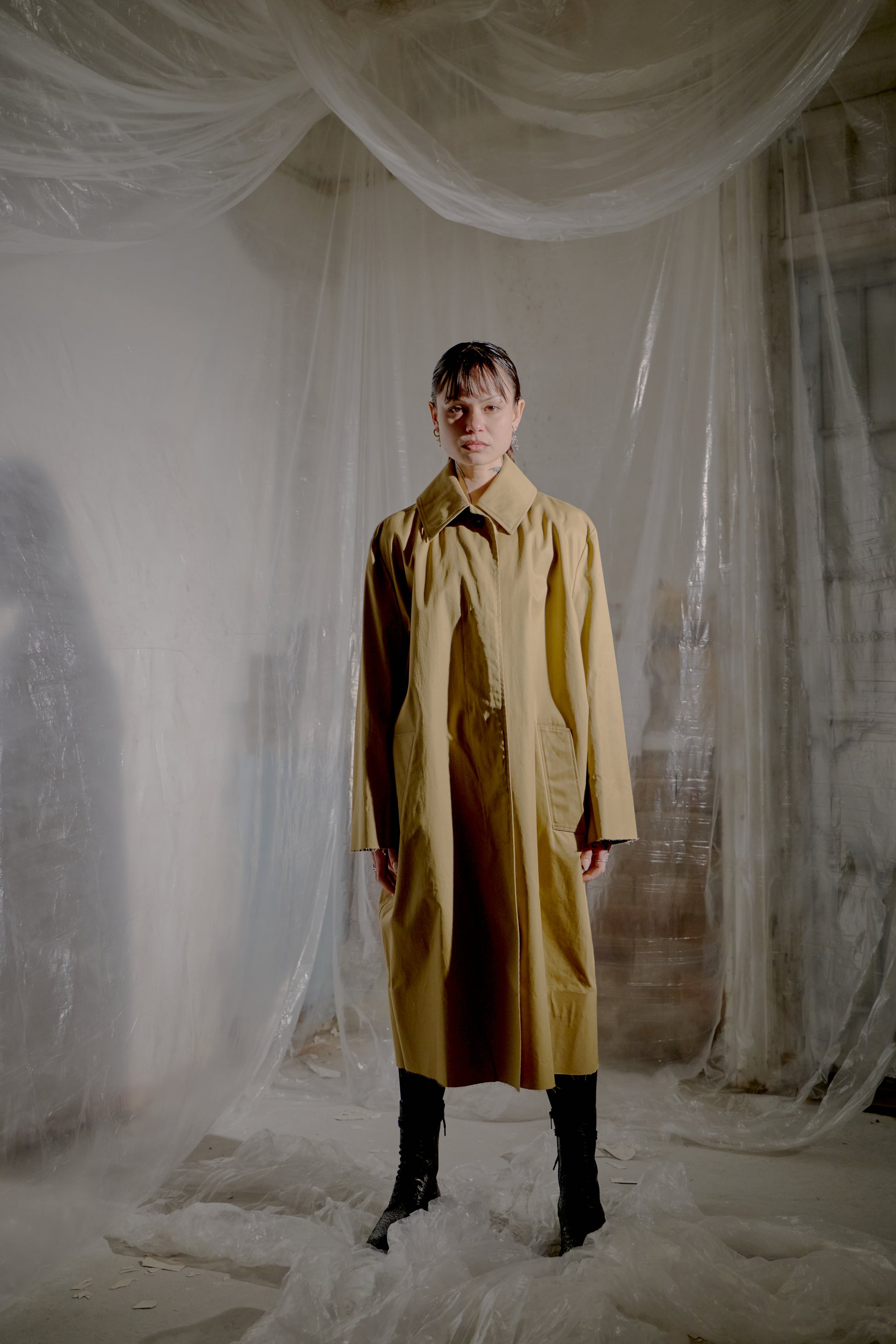 Person wearing a long brown Vetements coat standing in a room with white fabric draped around.