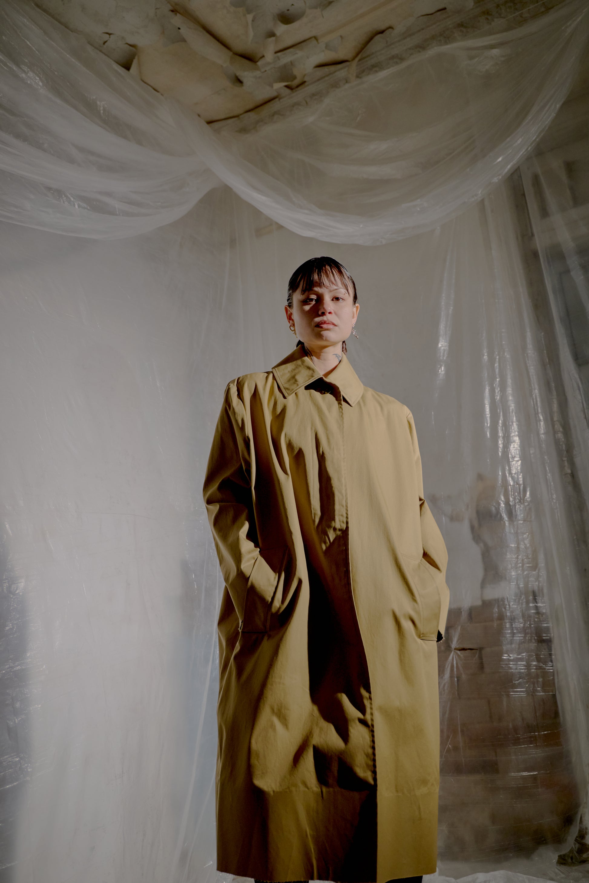 Person wearing a beige trench Vetements coat standing in a room with plastic sheeting on the walls.