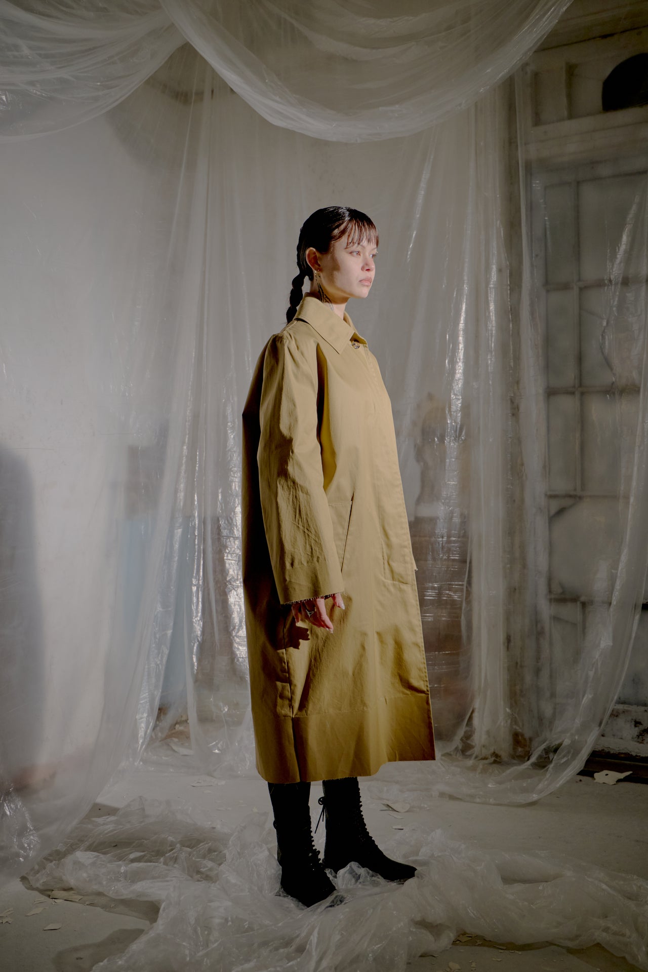 Person wearing a long brown Vetements coat standing in a room with white fabric draped around.