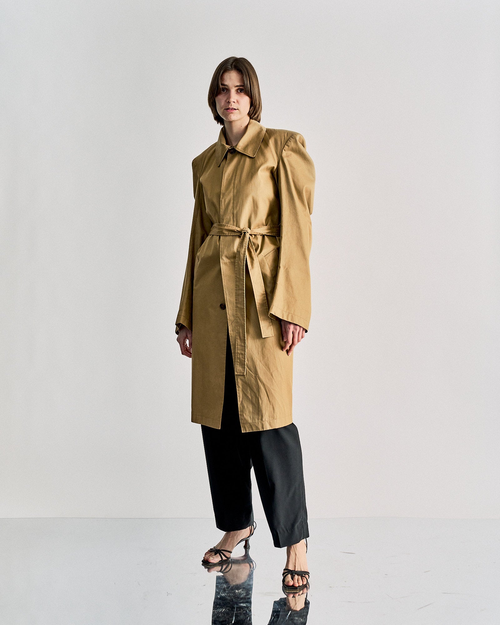 A person stands on a reflective floor in the Vetements SS 2016 Structured trench coat—a long tan piece with puffy shoulders—styled with black wide-leg pants and open-toed sandals, embodying Demna Gvasalia’s Spring Summer 2016 look against a plain white background.