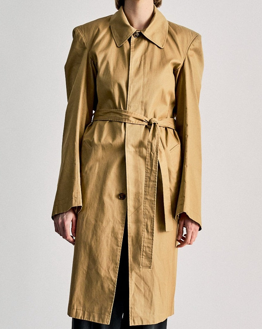 A person wears the SS 2016 Structured trench coat, a long tan Macintosh by Vetements, designed by Demna Gvasalia, with a belted waist, wide shoulders, and long sleeves, standing against a plain light background.
