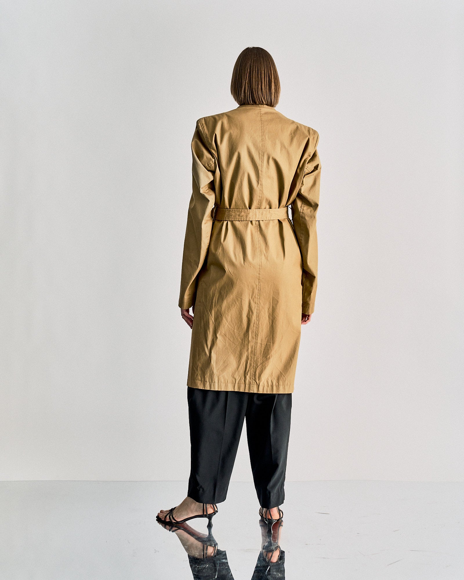 A person with short hair faces away from the camera, wearing a tan Vetements SS 2016 Structured trench coat, loose black pants, and black sandals against a plain light background.