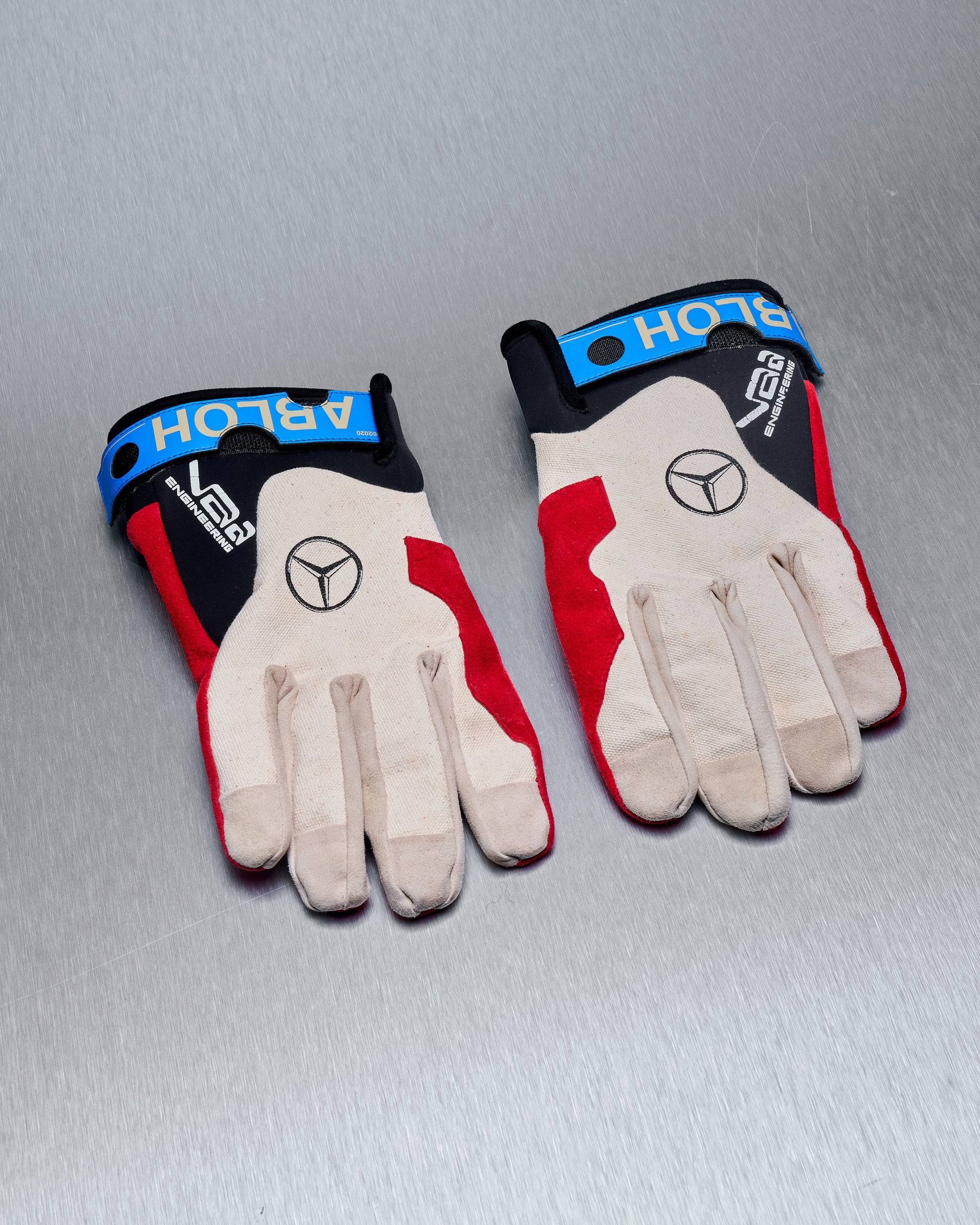 Two Virgil Abloh x Mercedes Benz racing gloves on a metallic surface. The gloves are beige with red and black accents, featuring blue straps labeled "ABLOH" and a black logo