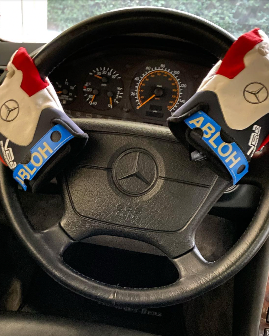 A close-up of a Mercedes-Benz steering wheel, adorned with two boxing gloves hanging over it. The gloves are white, red, and blue with the name "ABLOH." 