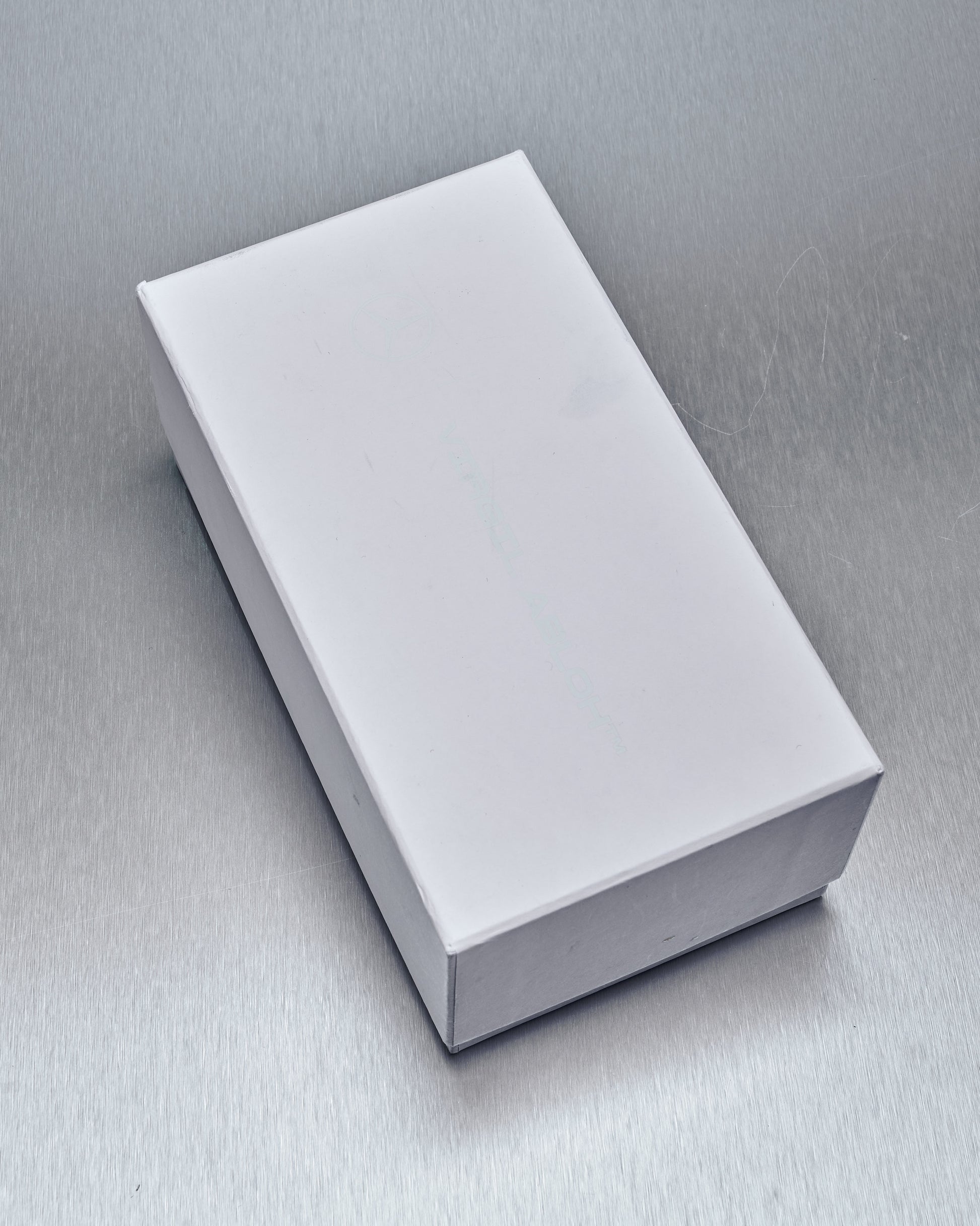 A white rectangular Virgil Abloh x Mercedes Benz box lies closed on a metallic surface. 