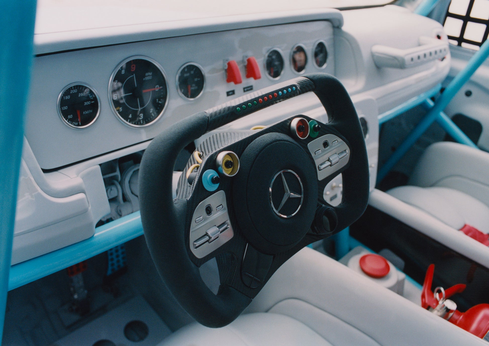 Close-up of a Mercedes-Benz race car interior, featuring a black steering wheel with coloured buttons, analog gauges, and a sleek, white dashboard.