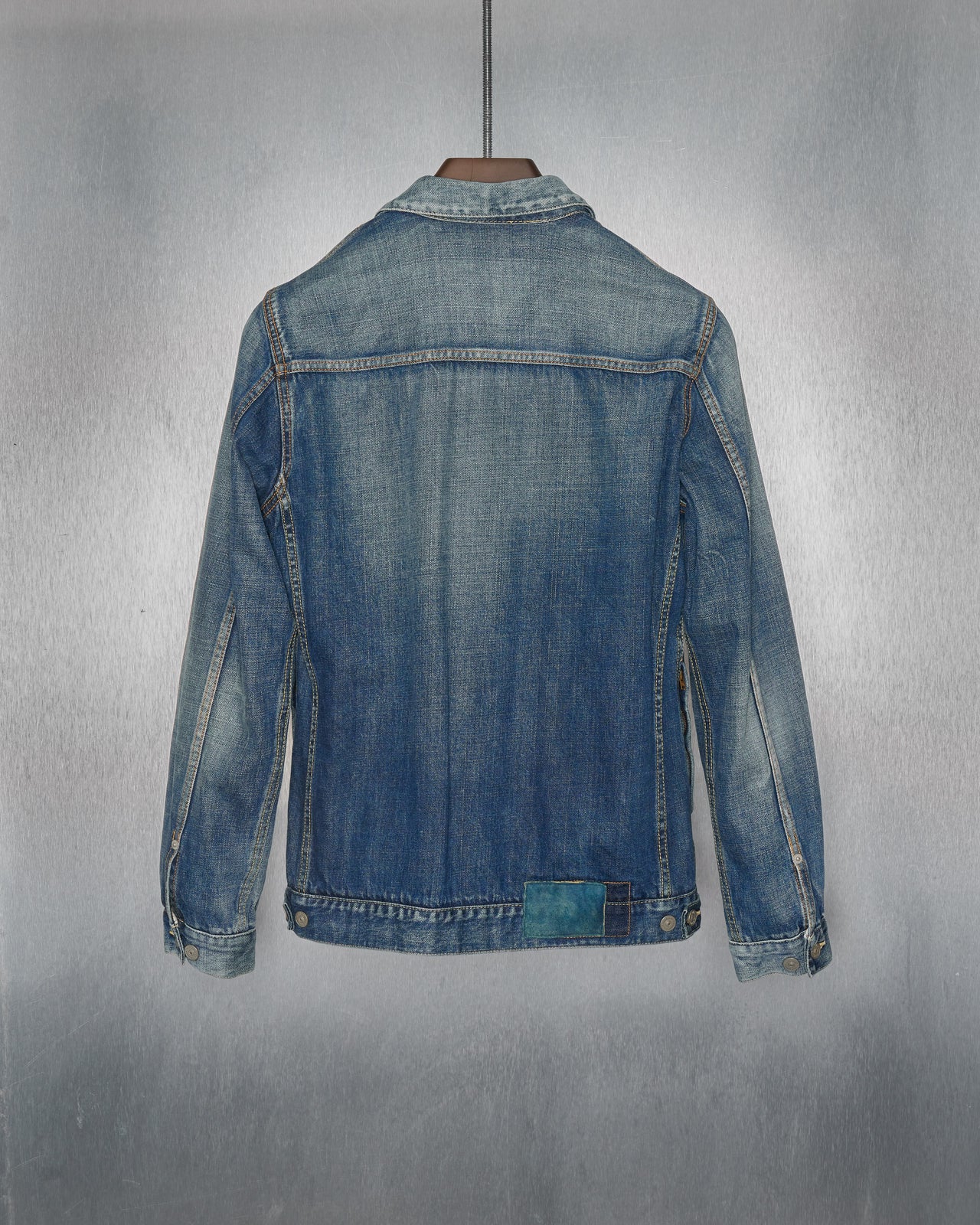 A blue denim Visvim jacket with a faded, worn-in look is hanging on a black hanger against a metallic textured background. The jacket has two chest pockets.