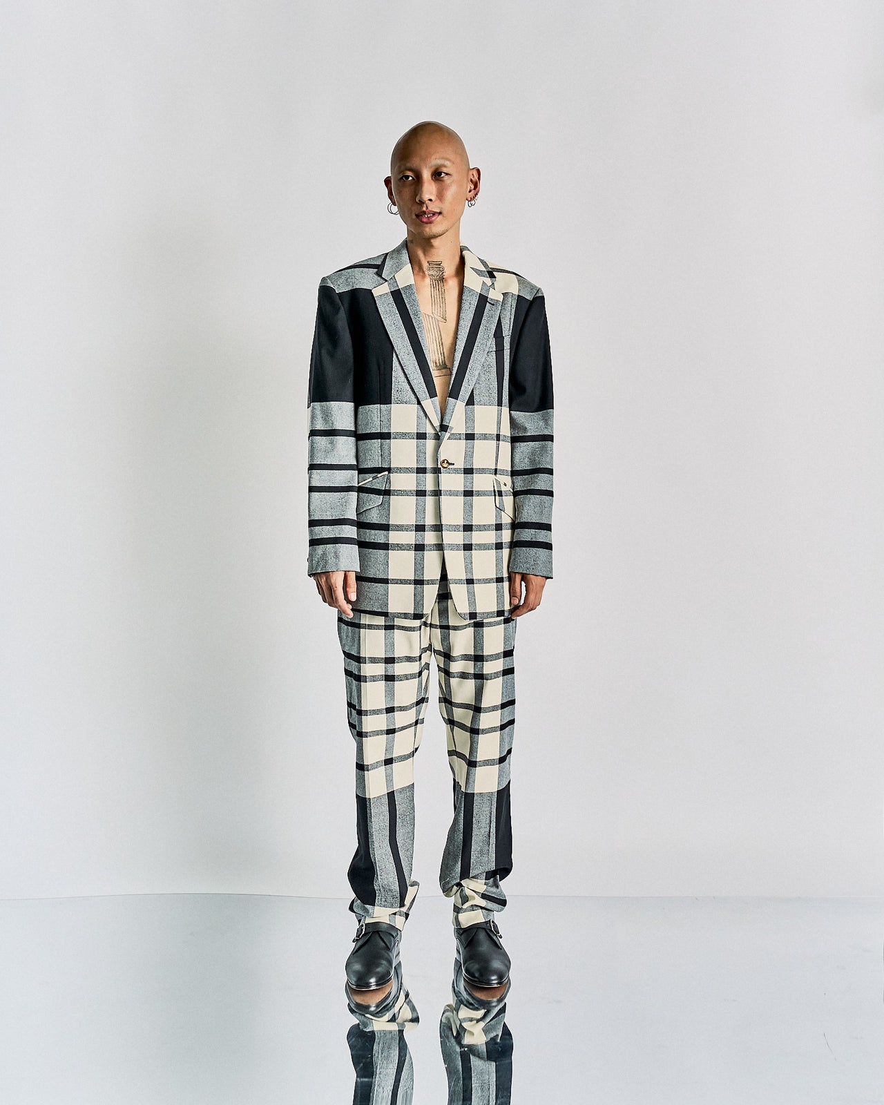 A person stands against a plain backdrop, wearing a checkered Vivienne Westwood suit with varying patterns in black and cream tones. The expression is neutral and relaxed.
