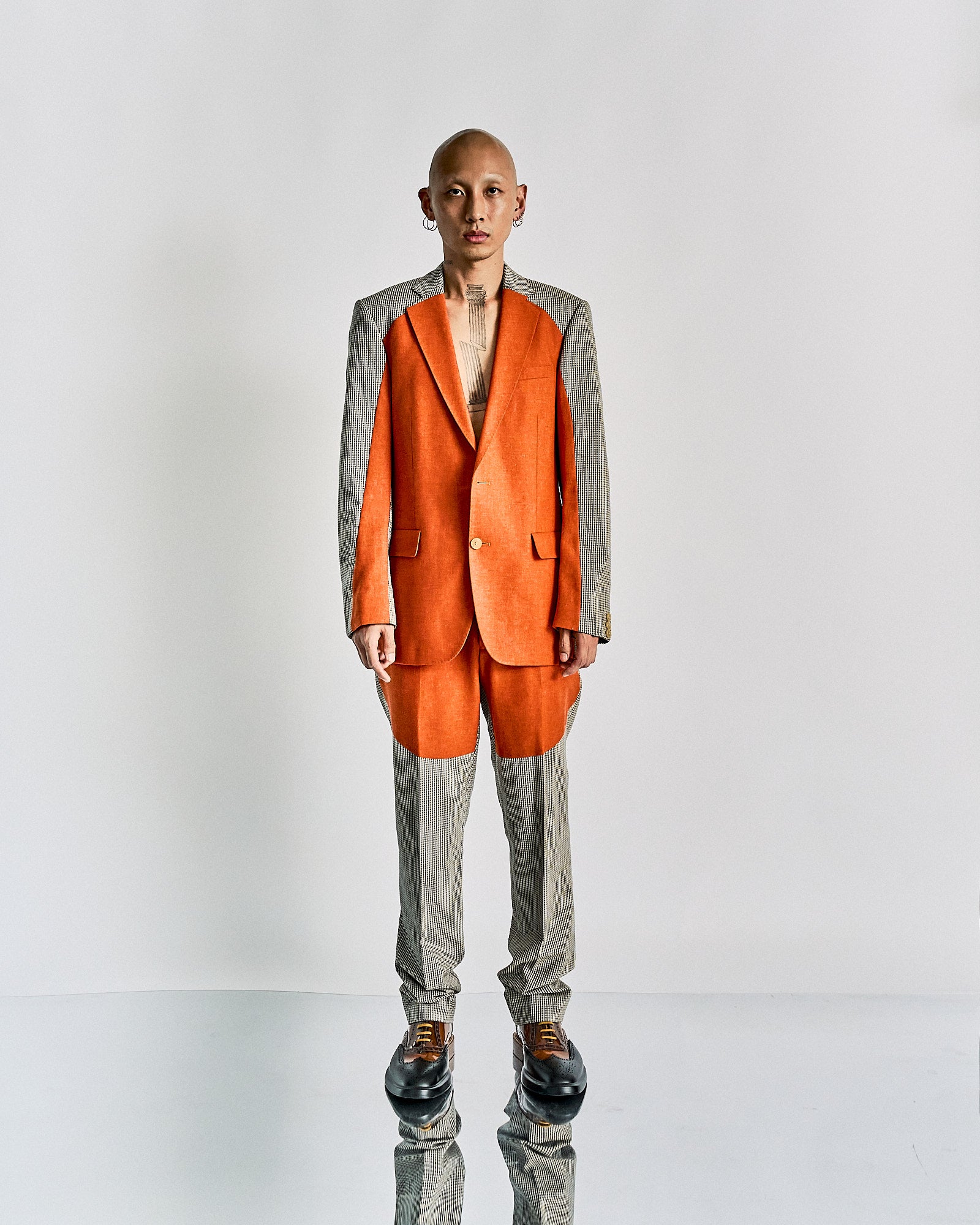 A person stands confidently in a Walter Van Beirendonck suit with an orange vest and pants along with grey, pinstriped sleeves and legs.