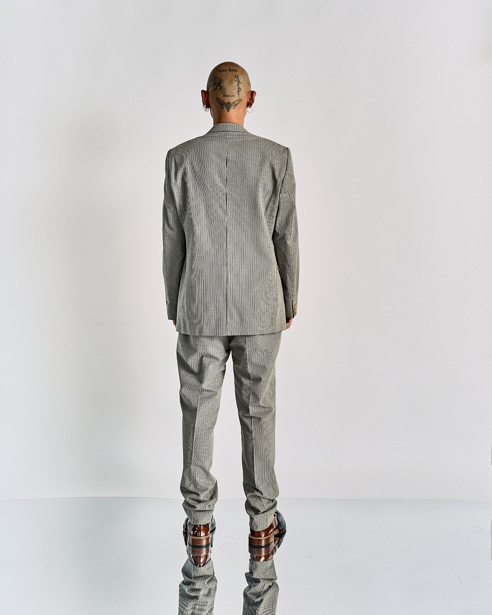 A person stands facing away, wearing a patterned grey Walter Van Beirendonck suit and brown shoes