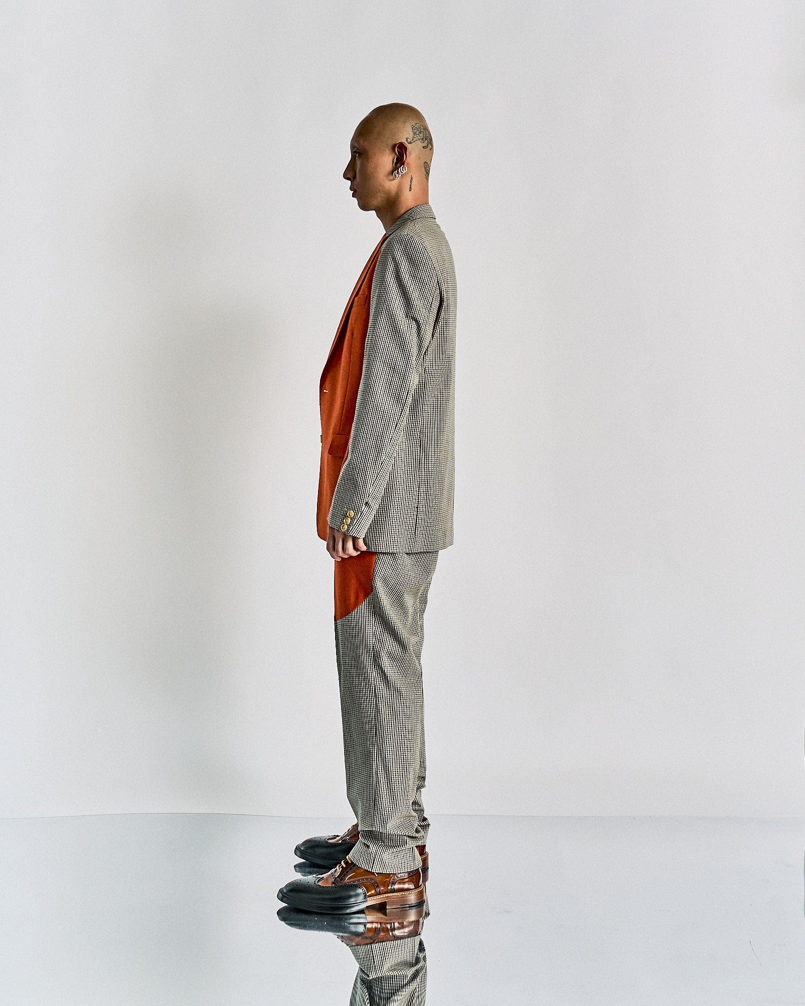A person in profile wears a grey plaid Walter Van Beirendonck suit with orange panels. They have a tattooed head, and the reflective floor mirrors their brown shoes.