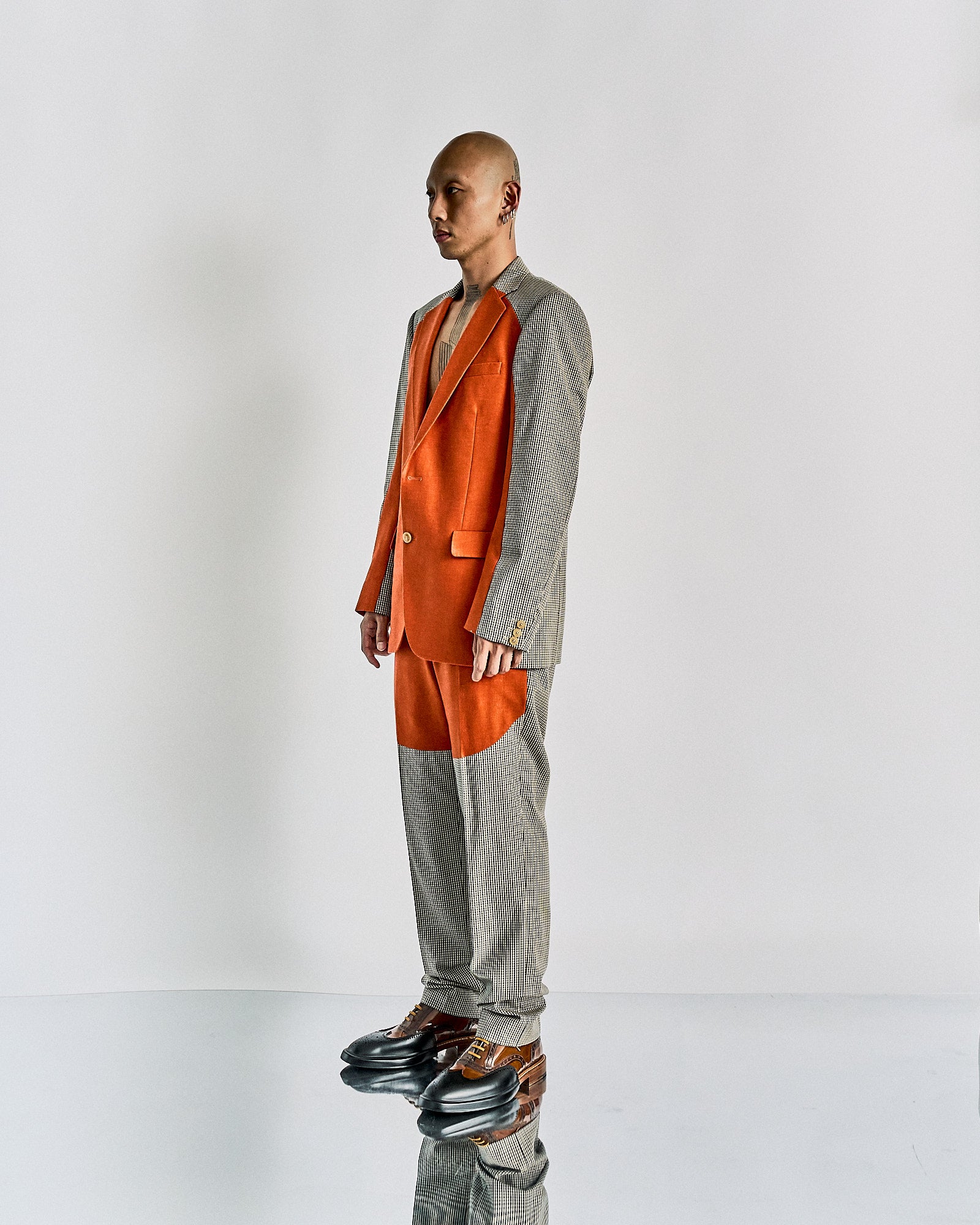 A person stands in a studio wearing a mixed-pattern Walter Van Beirendonck suit with grey and orange panels. 