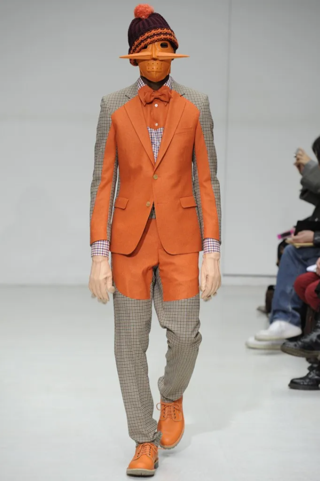 A person wearing a Walter Van Beirendonck two-piece suit with an orange circle motif. The suit is primarily gray with an orange circle on the chest and sleeves.