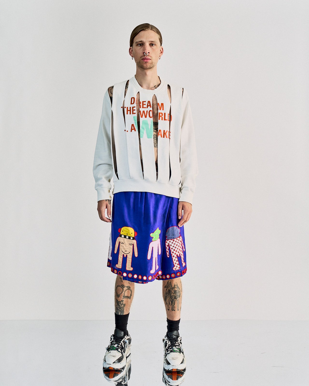 A model wearing a white Walter Van Beirendonck crewneck sweatshirt with the text 'DREAM THE WORLD AWAKE' on the front, alongside various diagonal slashes as a design feature. The model is also wearing colorful shorts with character illustrations.
