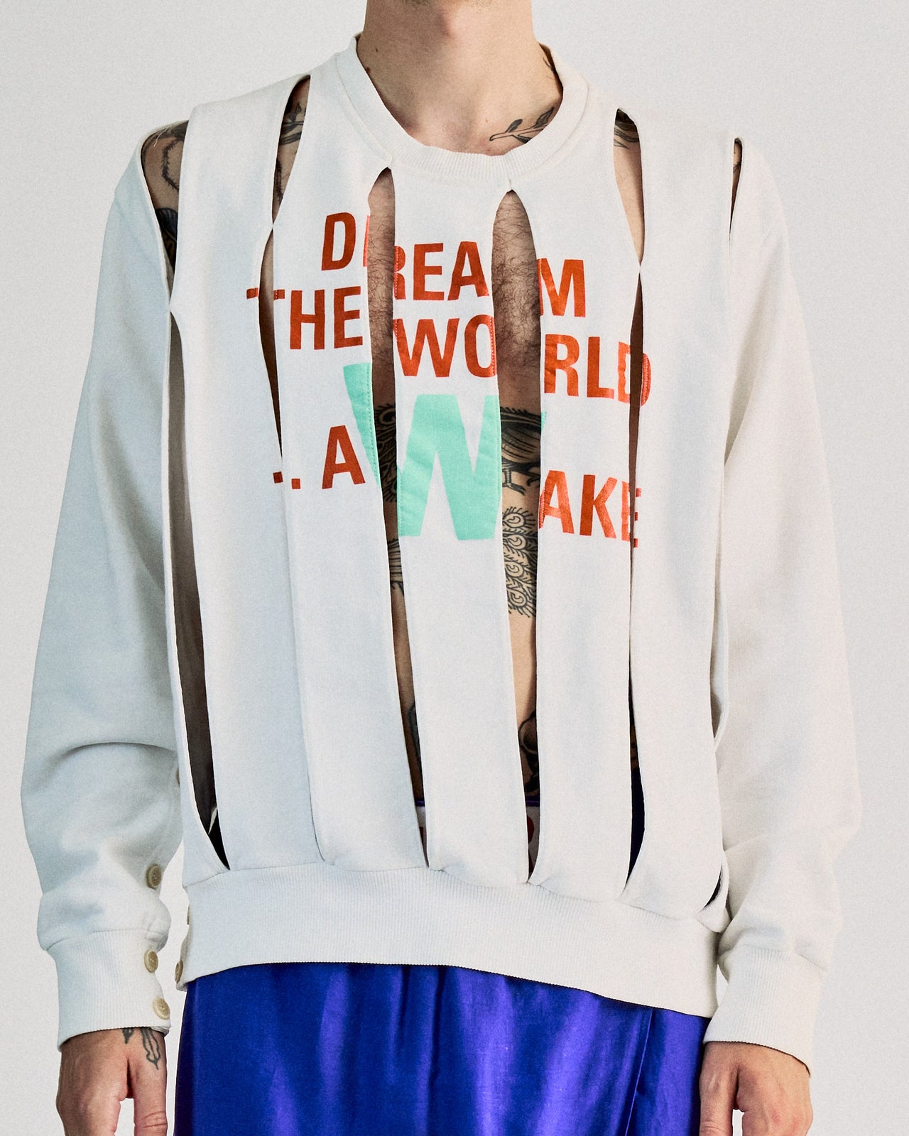 A male model wearing a white Walter Van Beirendonck crewneck sweatshirt with orange and green shorts, and face paint resembling a shaved head with the text 'DREAM THE WORLD AWAKE' on the sweatshirt.