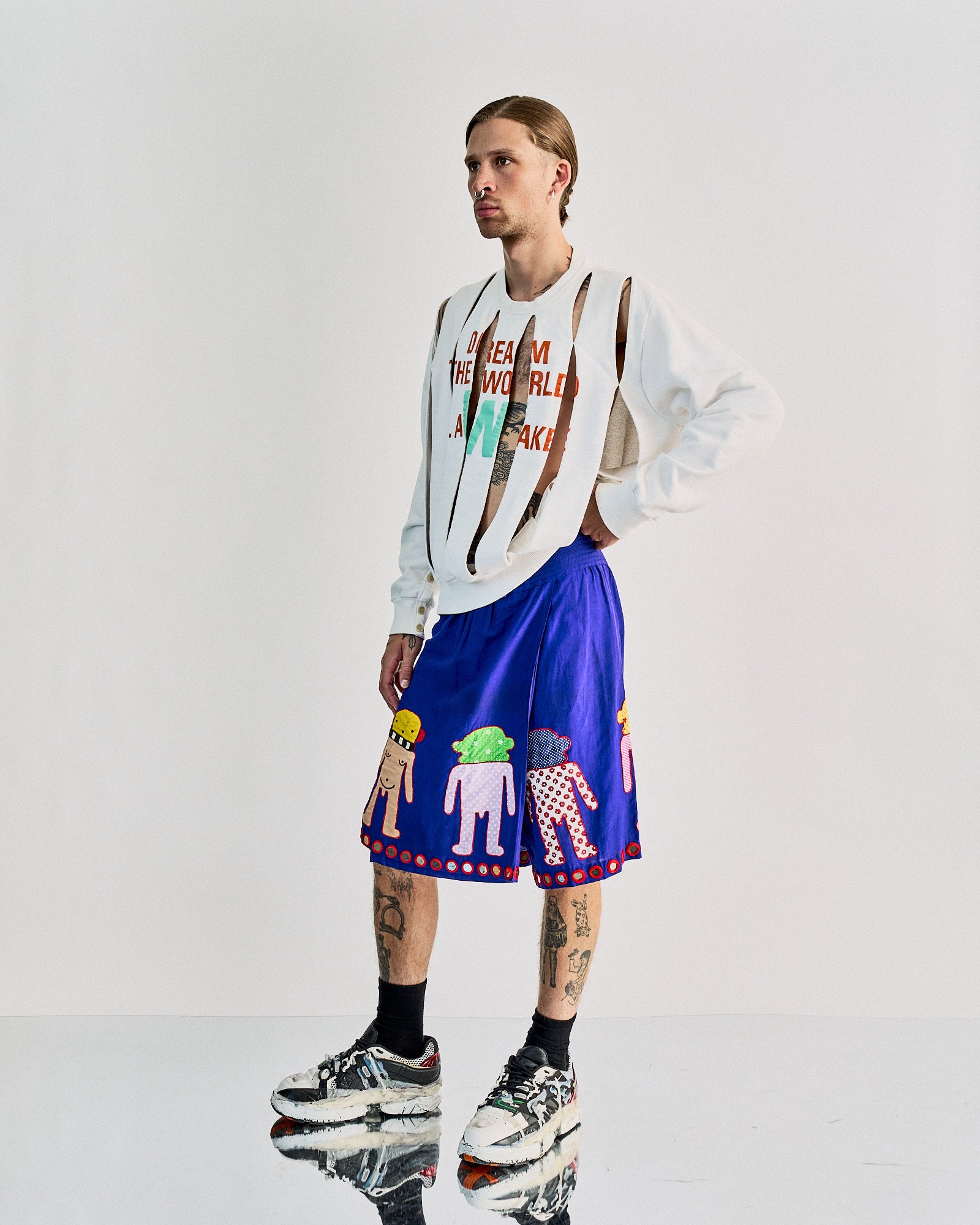 A person with tattoos wears a distressed white Walter Van Beirendonck  sweater and colorful shorts featuring cartoon figures. They stand on a reflective surface. Edgy and artistic vibe.