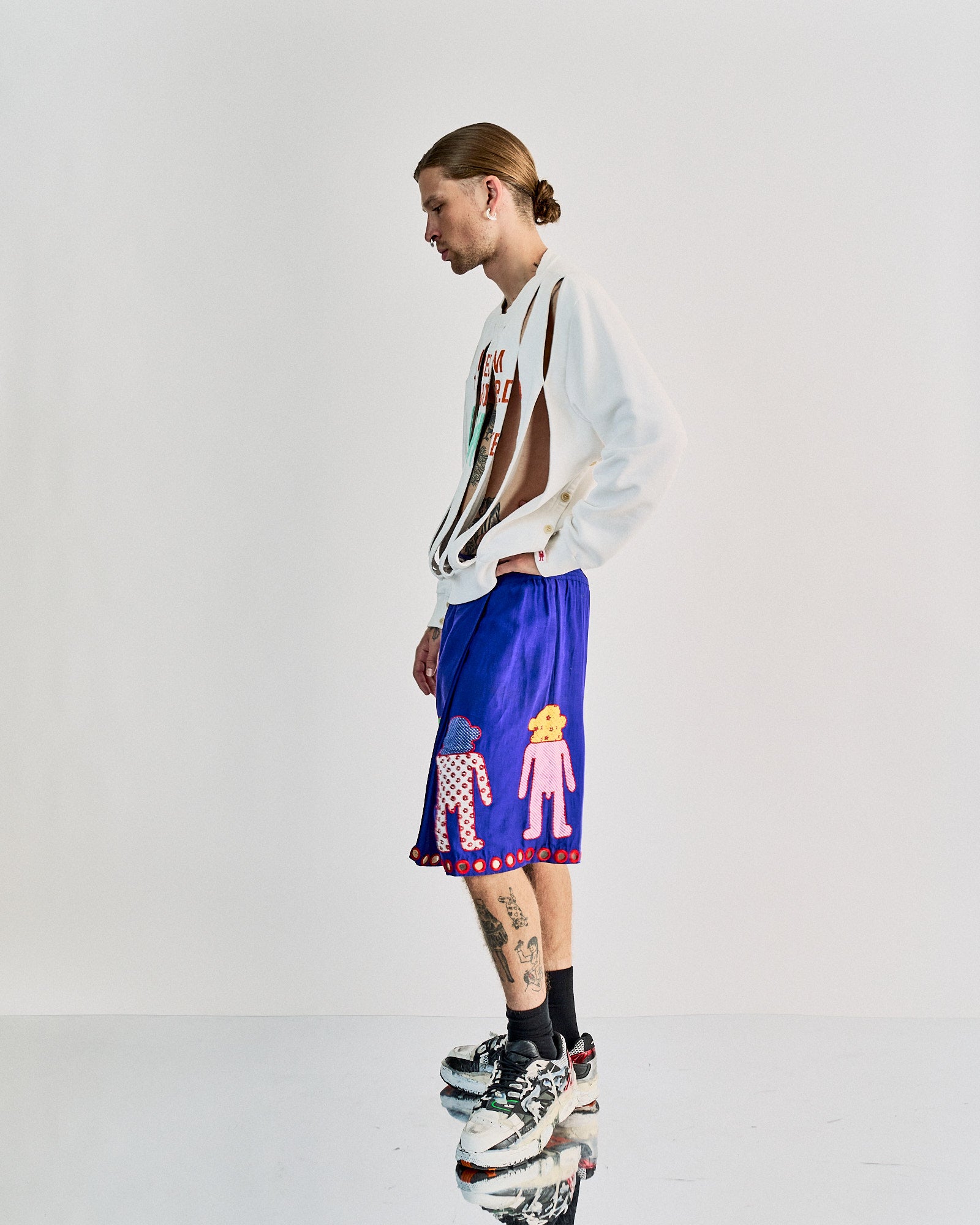 A person in a slashed white Walter Van Beirendonck  sweatshirt reads "I Dream The World Awake," paired with purple shorts featuring playful, colorful characters. They wear chunky sneakers, standing pensively, suggesting a trendy, artistic vibe.
