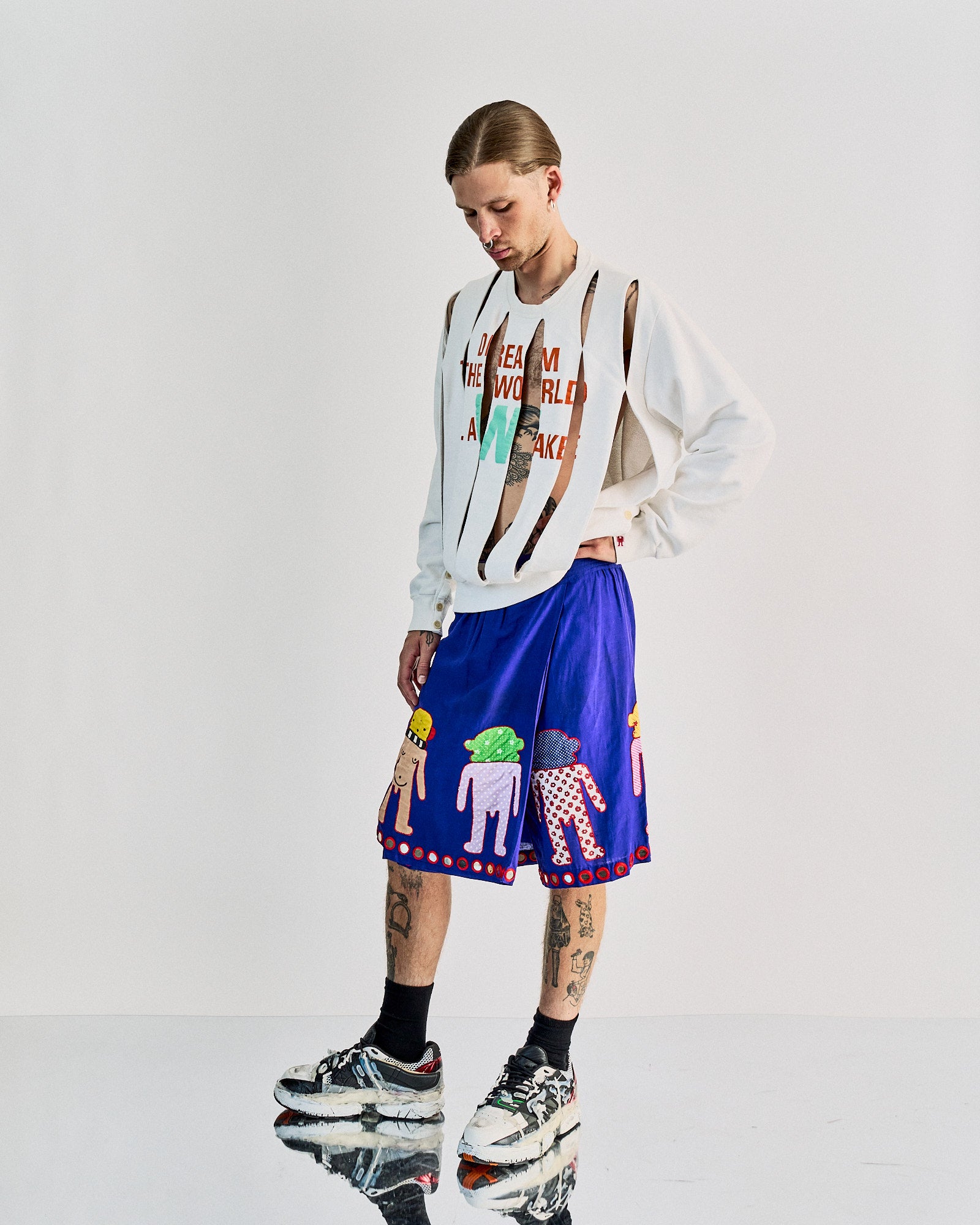 A person in a slashed white Walter Van Beirendonck  sweatshirt reads "I Dream The World Awake," paired with purple shorts featuring playful, colorful characters. They wear chunky sneakers, standing pensively, suggesting a trendy, artistic vibe.