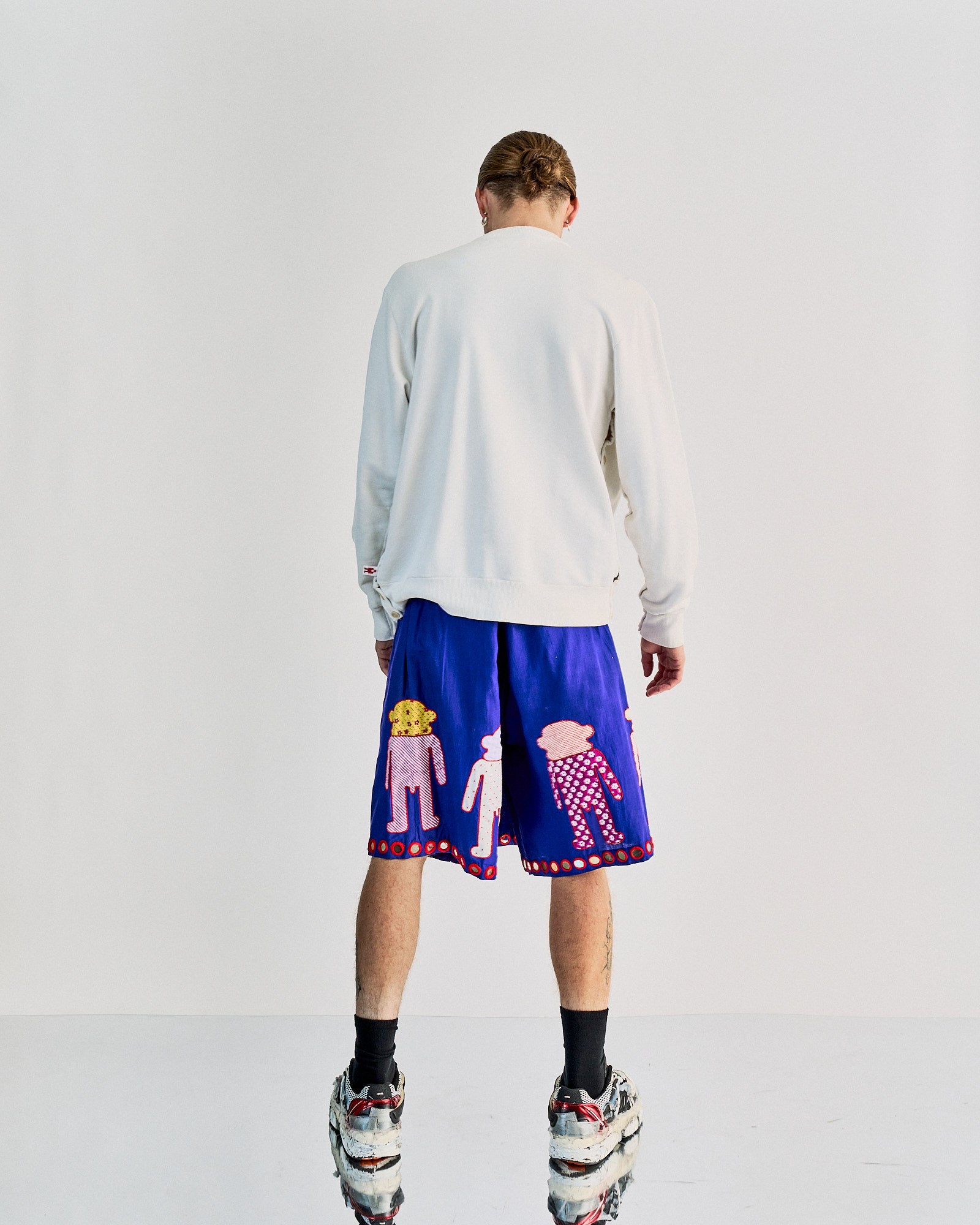 A person stands with their back to the camera, wearing a white Walter Van Beirendonck top and vibrant blue shorts with playful cartoon figures. They have sneakers and black socks.