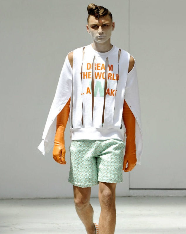 A male model wearing a white Walter Van Beirendonck crewneck sweatshirt with orange and green shorts, and face paint resembling a shaved head with the text 'DREAM THE WORLD AWAKE' on the sweatshirt.