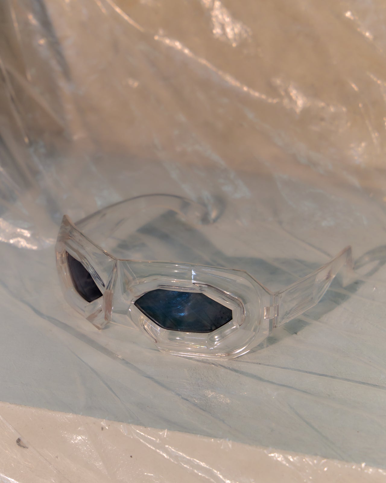 Clear Walter Van Beirendonck x Linda Farrow sunglasses with dark lenses on a reflective surface