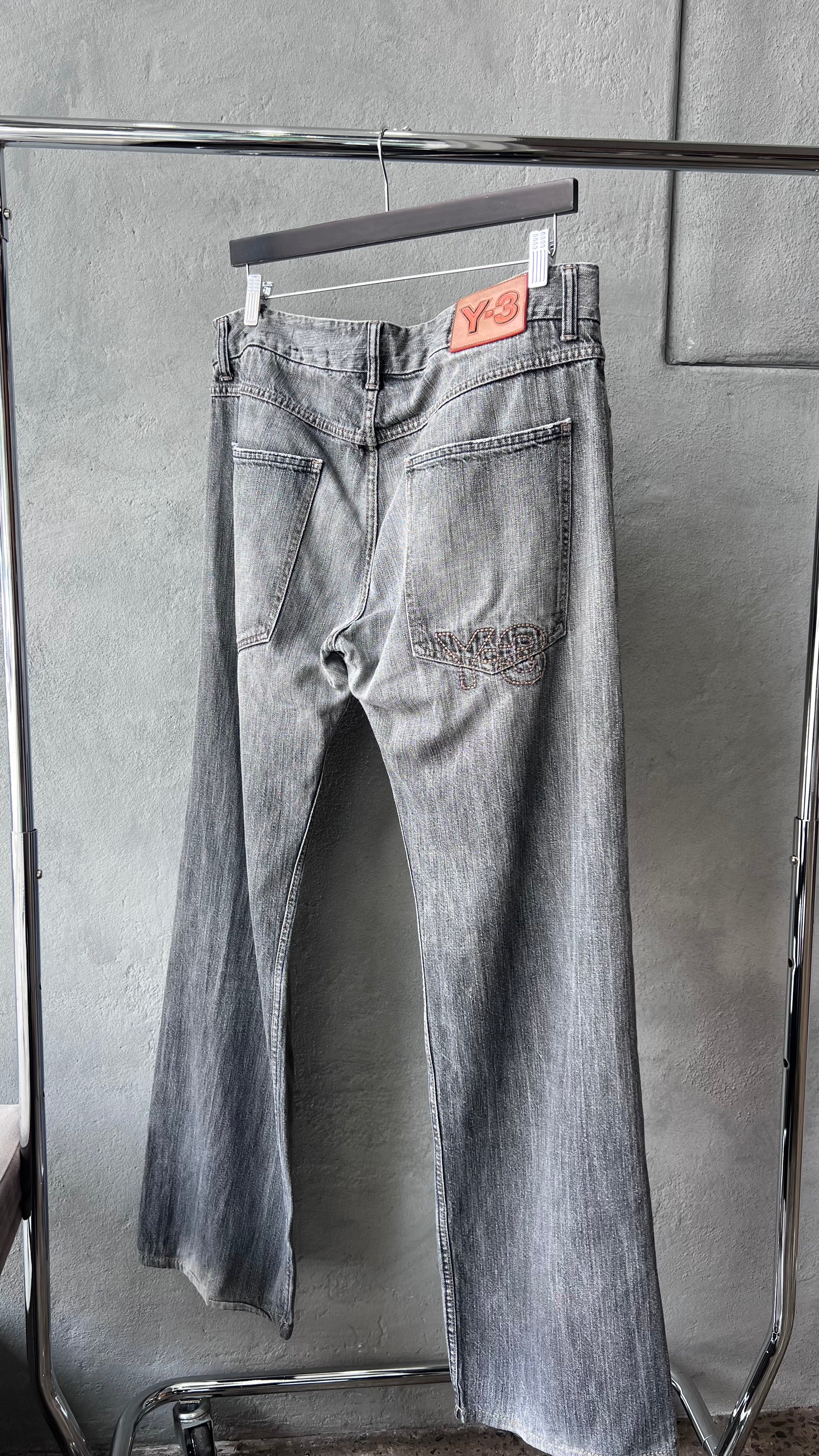 Pair of light grey Y-3 jeans hanging on a rack against a gray wall.