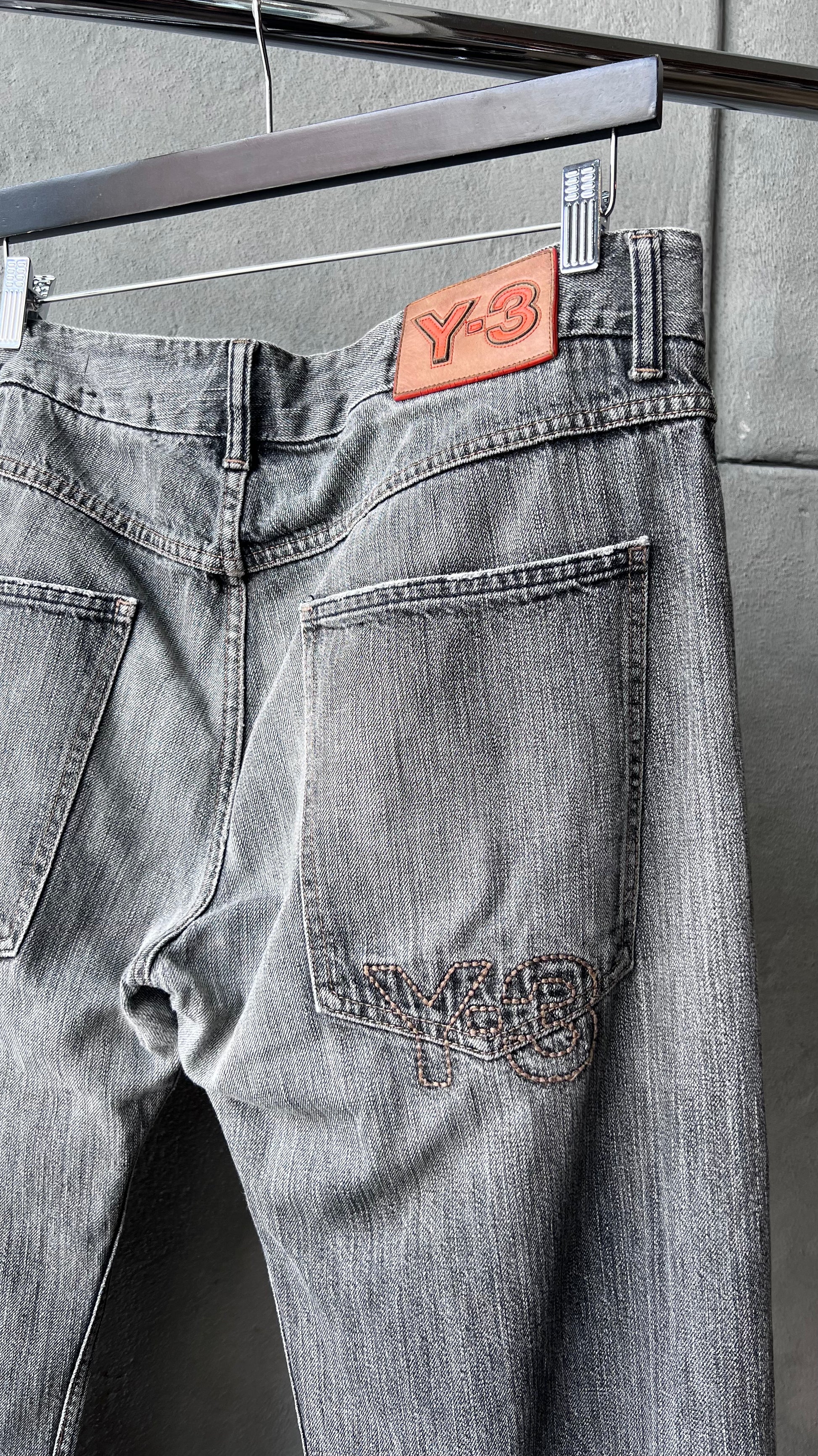 Gray Y-3 jeans with a brand patch on a hanger against a gray wall.