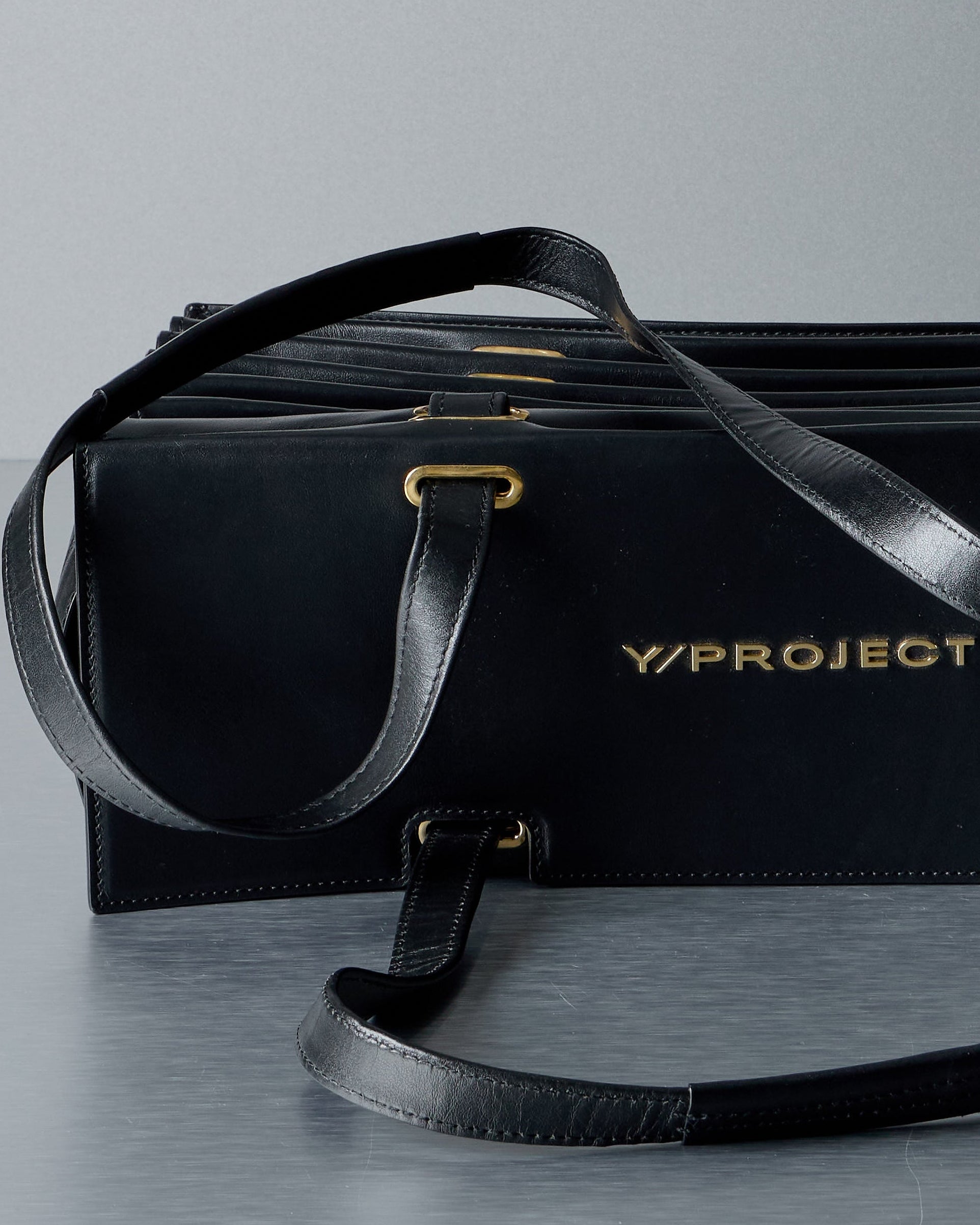 Black leather bag with gold 'Y/Project' logo on a gray background