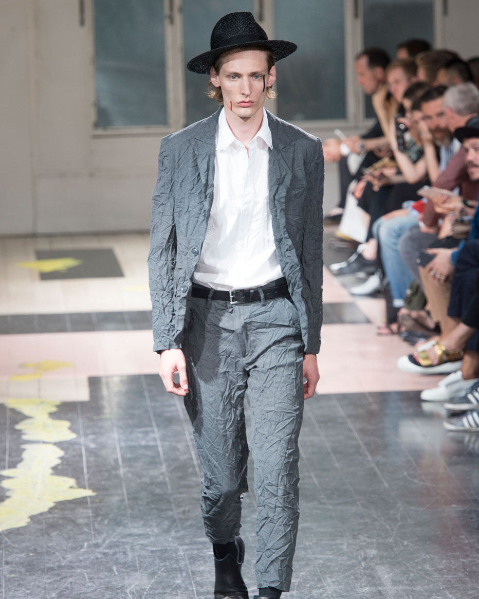 Man in a gray Yohji Yamamoto suit and black hat walking on a runway with an audience.