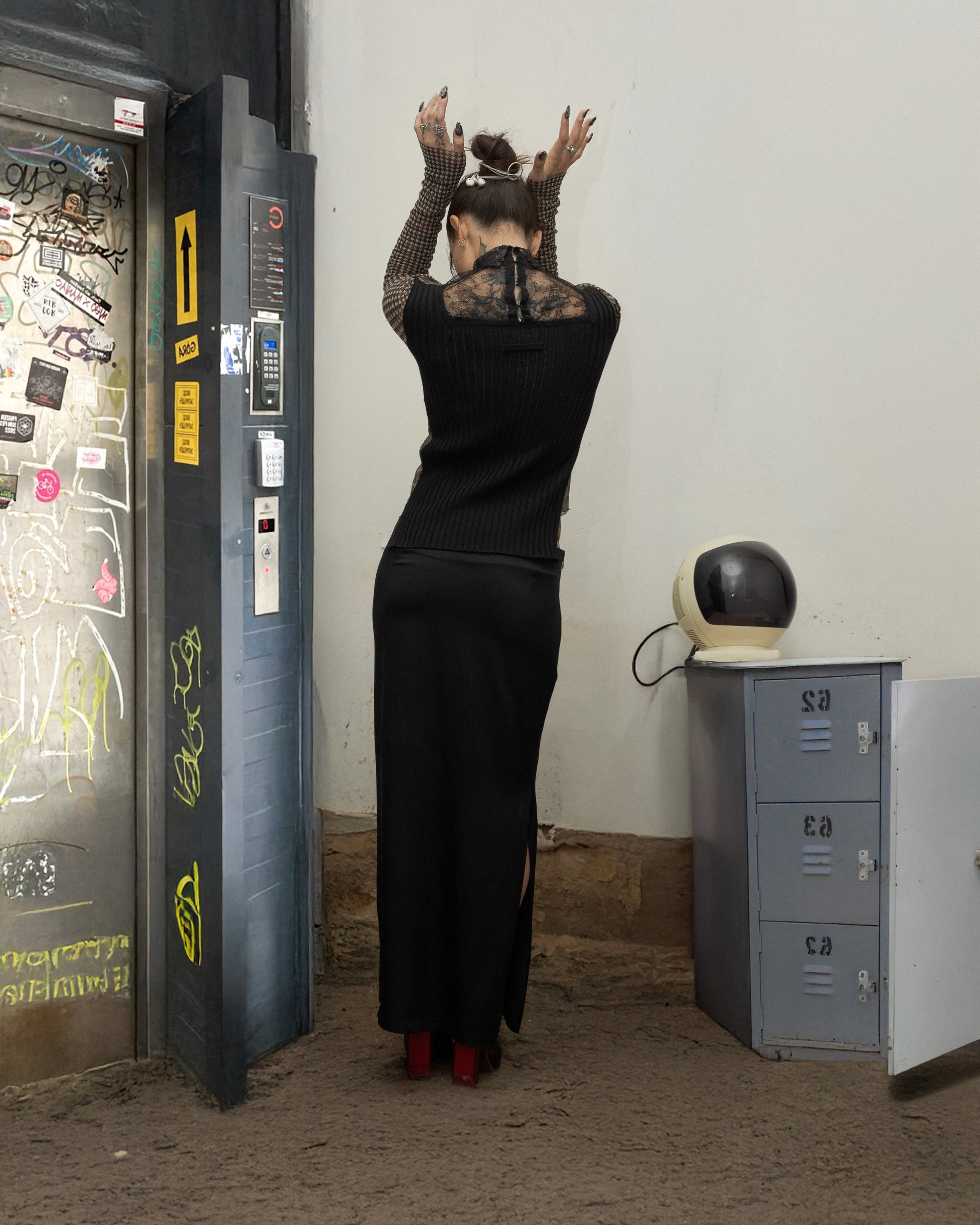 Person in a long black dress standing in front of a payphone booth with a white wall and locker in the background.
