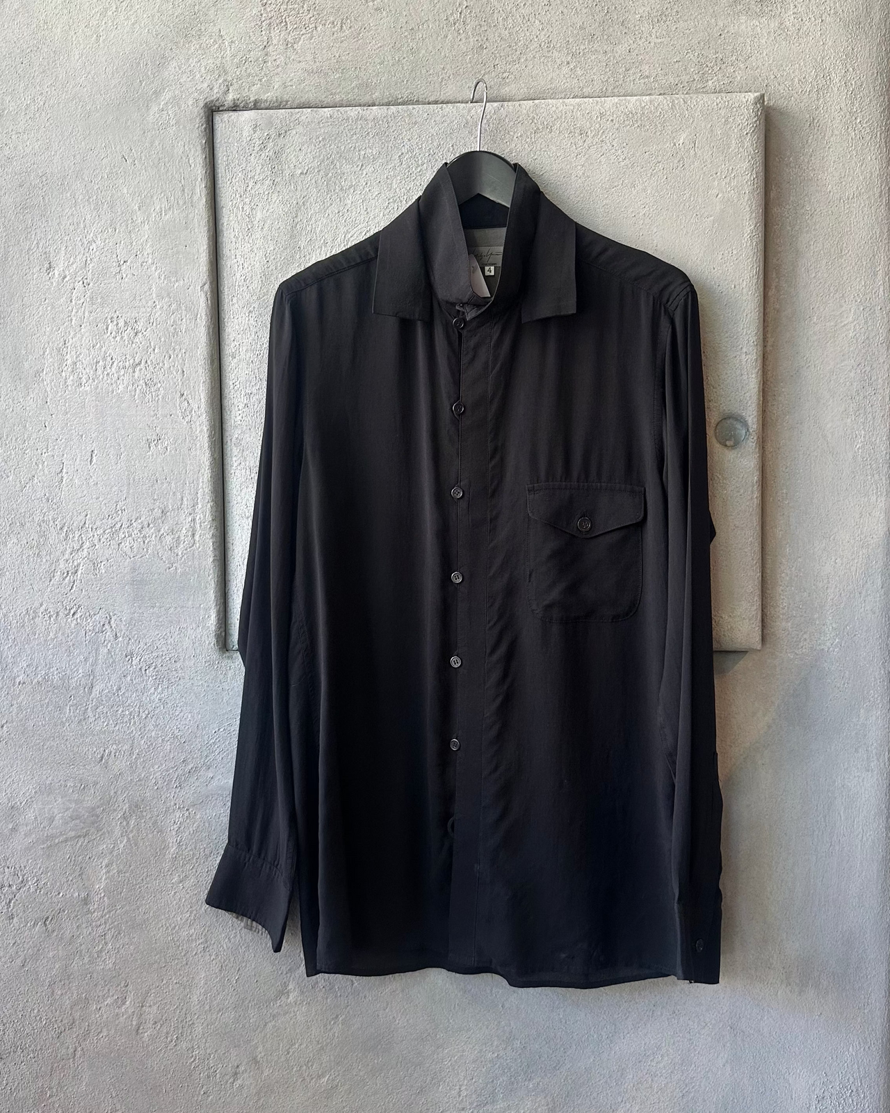 Black Yohji Yamamoto shirt hanging on a hook against a textured wall