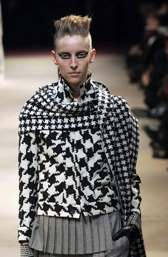 Model on runway wearing a bold black and white houndstooth-patterned outfit from Yohji Yamamoto with sharp makeup and a high mohawk.