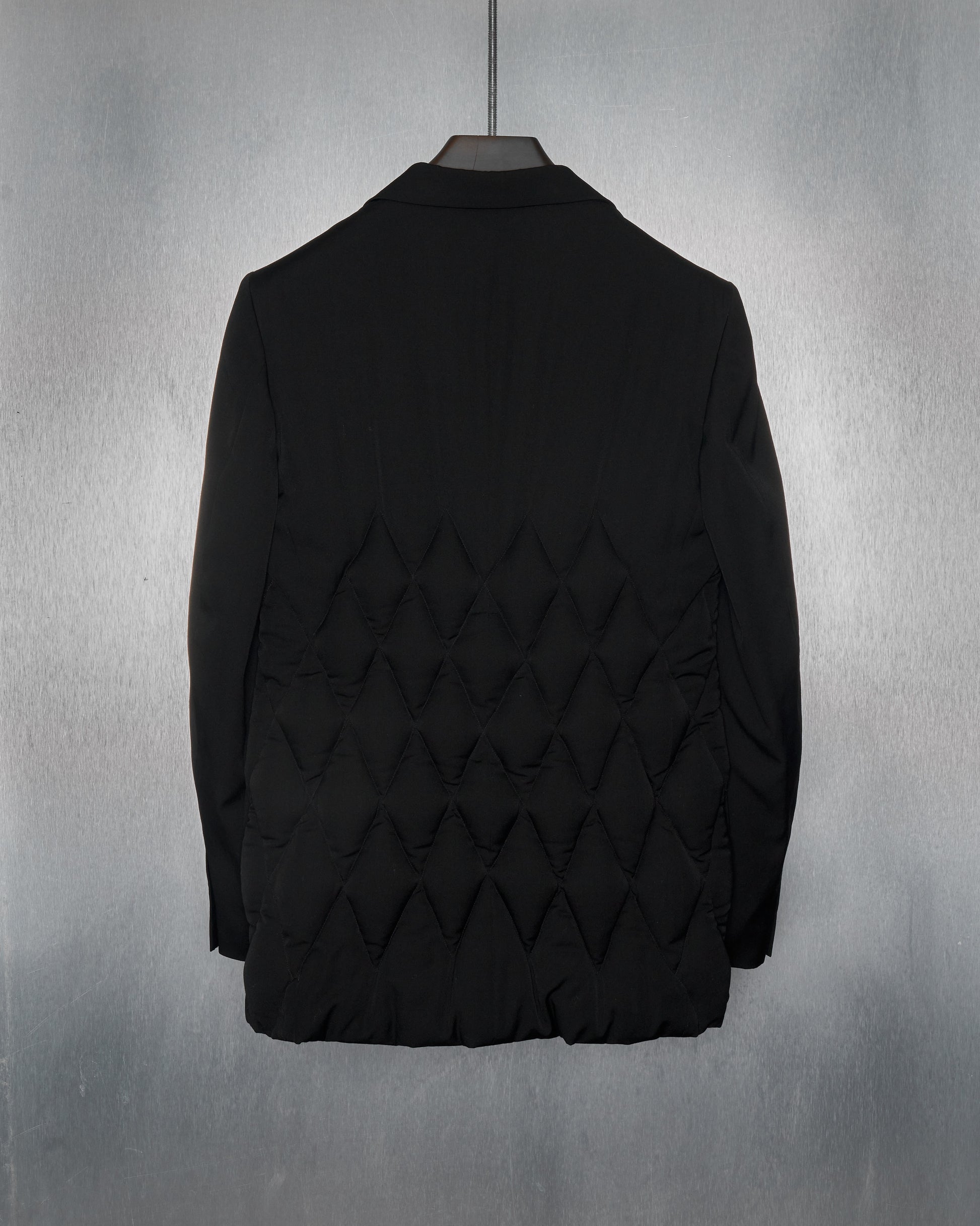 A black Yohji Yamamoto jacket with a quilted diamond pattern on the back hangs against a textured, metallic gray background, conveying a sleek and modern style.