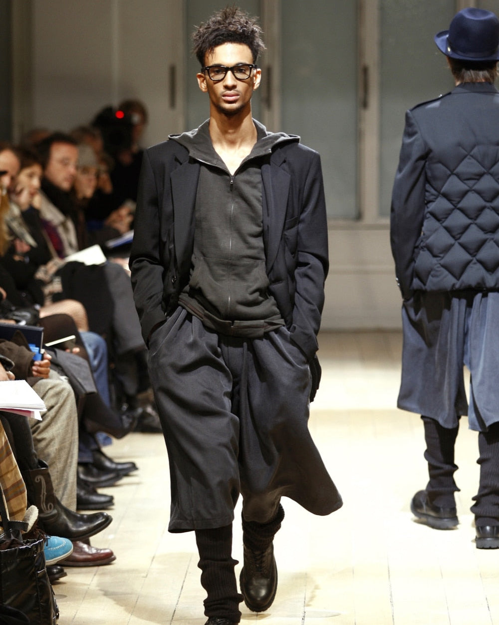 A model confidently walks down a Yohji Yamamoto fashion runway in a stylish dark outfit. Seated audience members observe attentively, enhancing the event's sophistication.