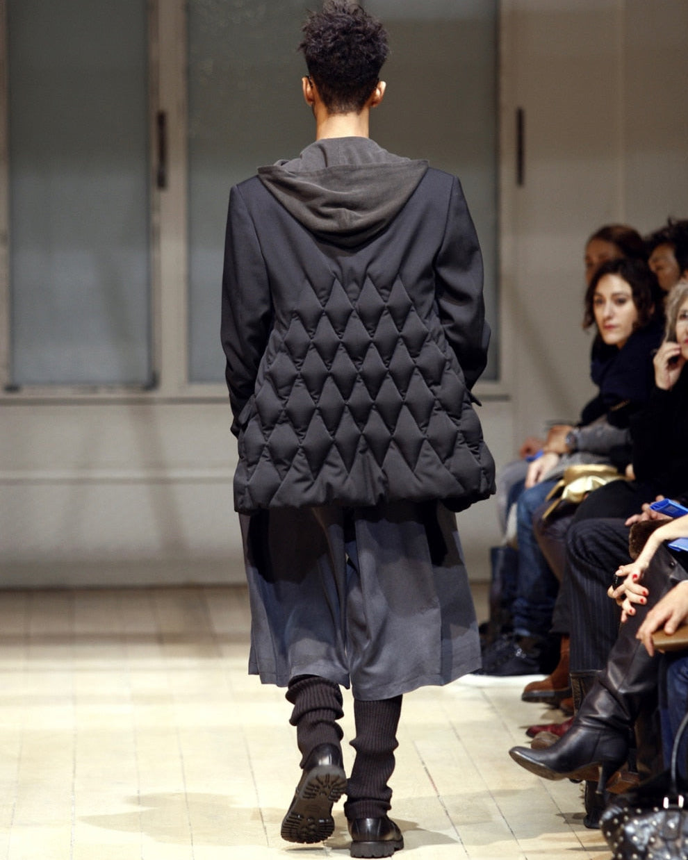 A model walks down a wooden runway wearing a black quilted Yohji Yamamoto coat layered over a gray outfit. Audience members sit on either side, watching intently.