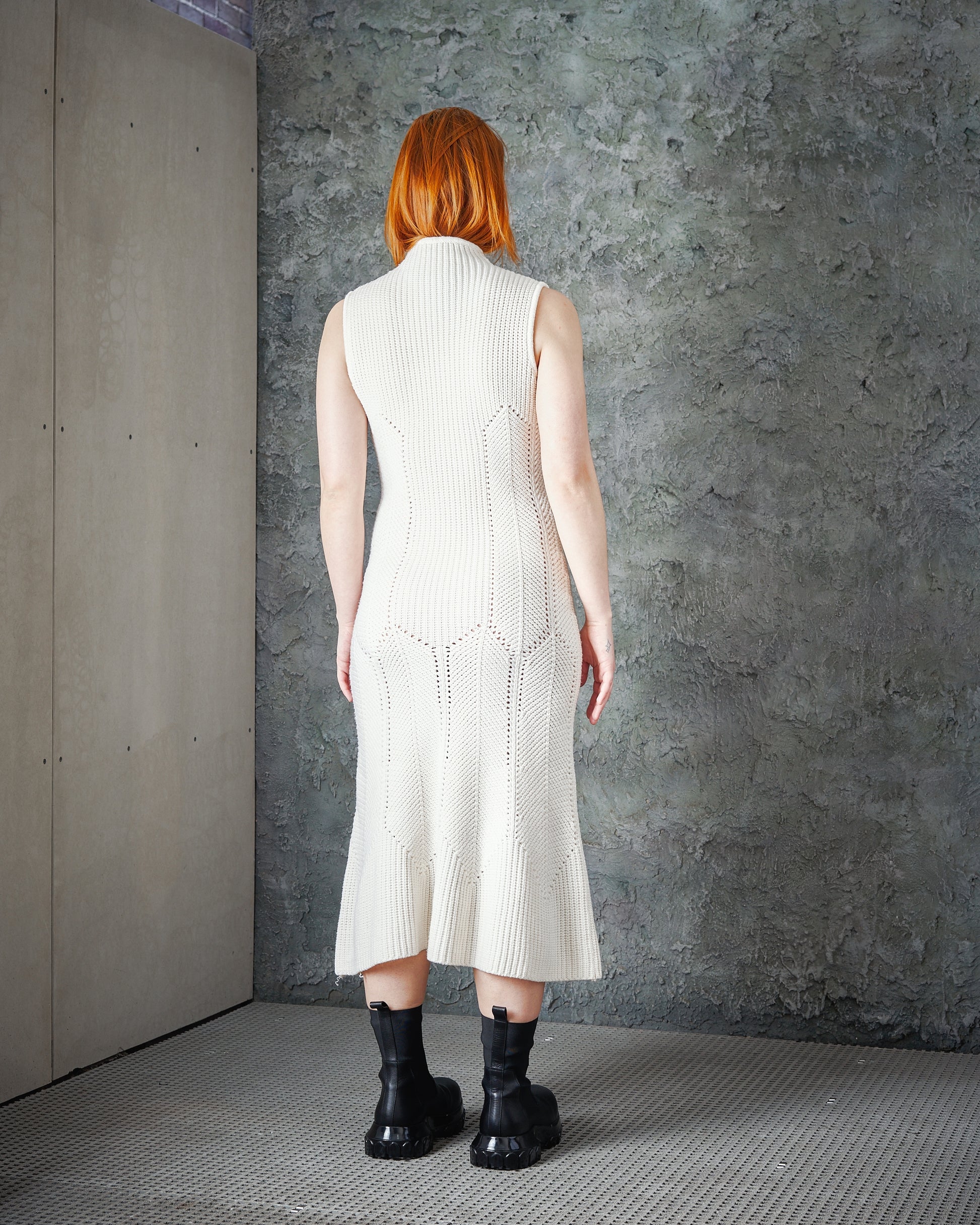 A person with red hair stands facing a textured grey wall, wearing a sleeveless cream knit Alexander McQueen dress and black boots.