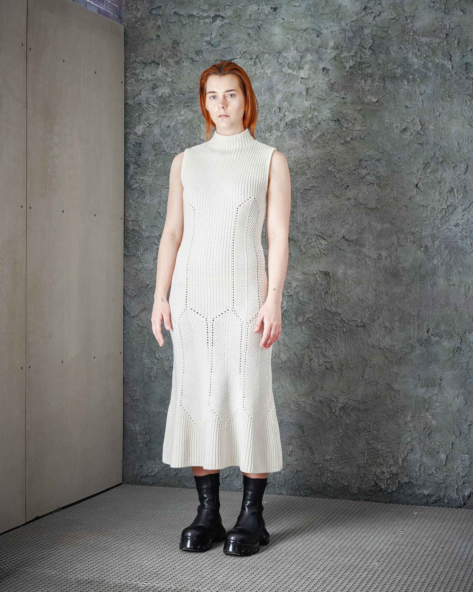 A person with short hair stands against a textured grey wall, wearing a sleeveless, high-neck, cream knit Alexander McQueen dress and black chunky boots.