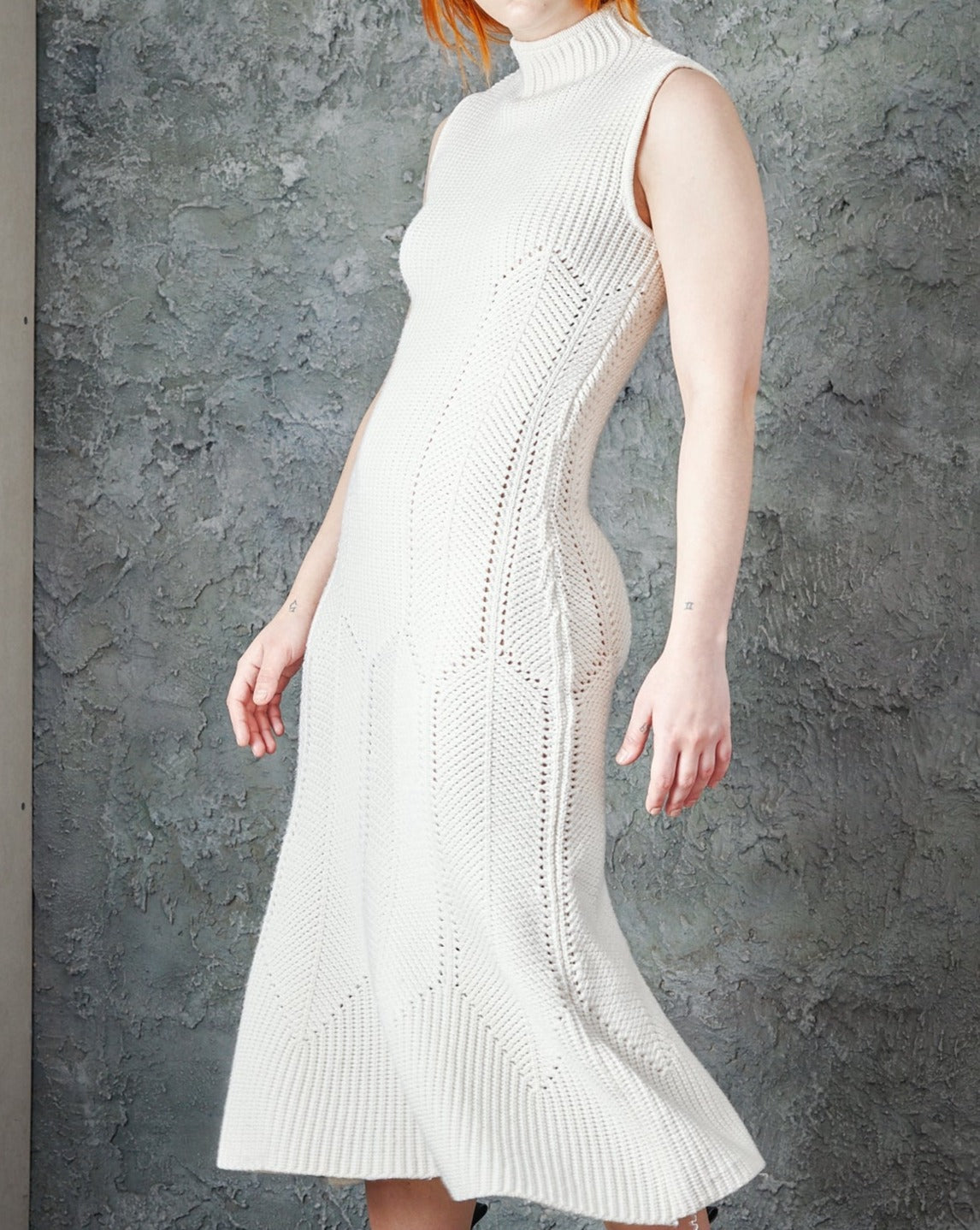 Woman in a sleeveless, textured white knit Alexander McQueen dress stands against a textured grey wall. 