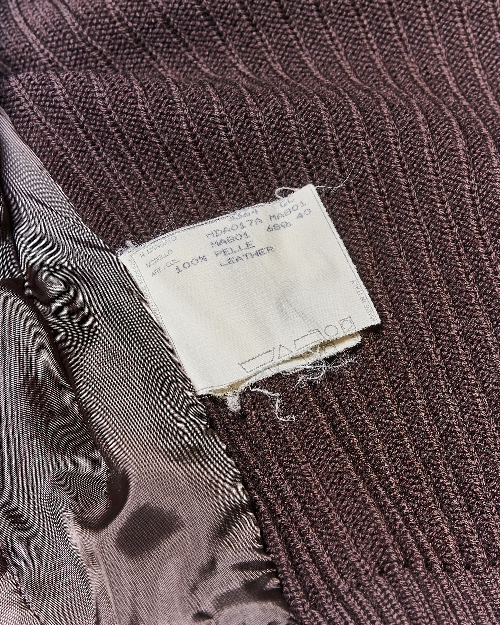 Close-up of a brown ribbed knit fabric with a white label showing care instructions for Alexander McQueen. Label includes text about material composition, mentioning "100% leather."
