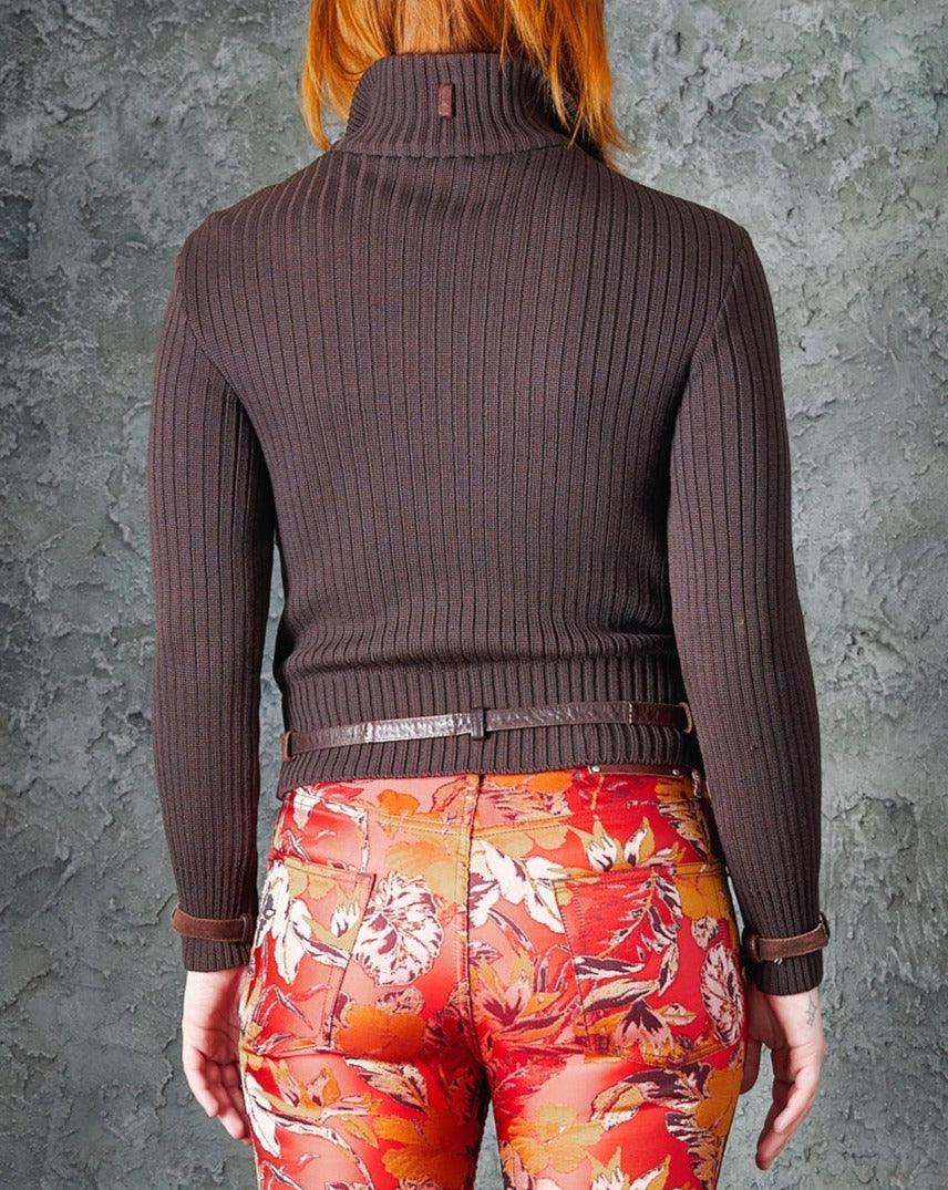 A person with red hair is seen from the back, wearing a dark brown Alexander McQueen ribbed turtleneck jacket and vibrant red floral pants. 