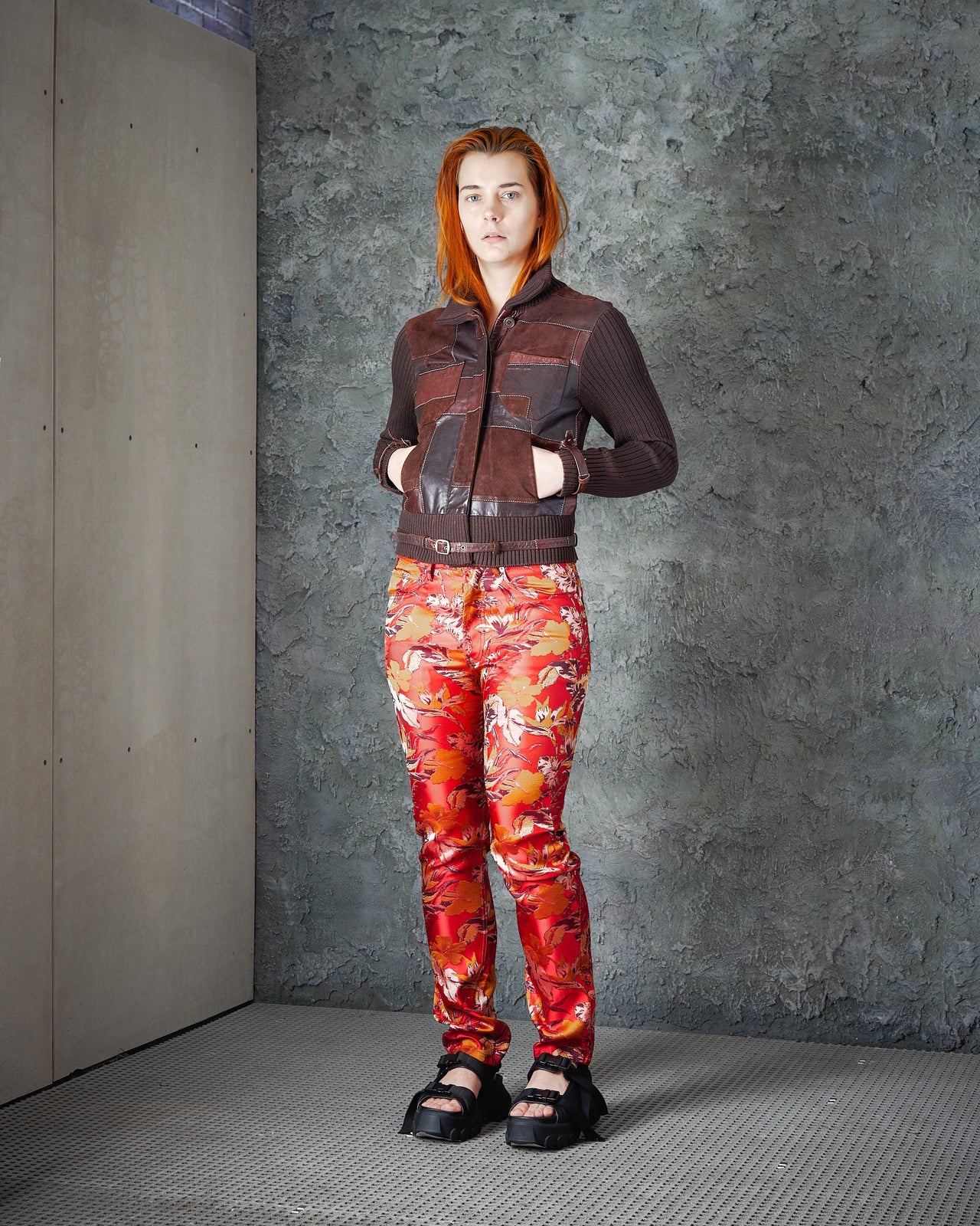 A woman standing in a runway setting, wearing an Alexander Mcqueen FW 2000 Look 27 Patchwork Leather Knit Jacket in red and orange hues with a ribbed knit collar and sleeves, featuring detachable leather belts.