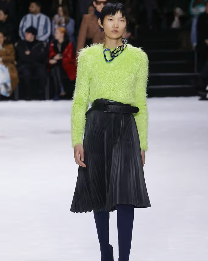 Model wearing a bright green Balenciaga sweater and black pleated skirt on a runway.